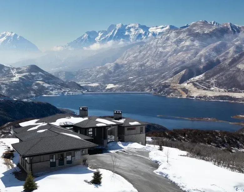 Aerial view of a modern mountain home with a dark shingled roof and stone siding, snow-covered terrain, and a lake and mountain backdrop