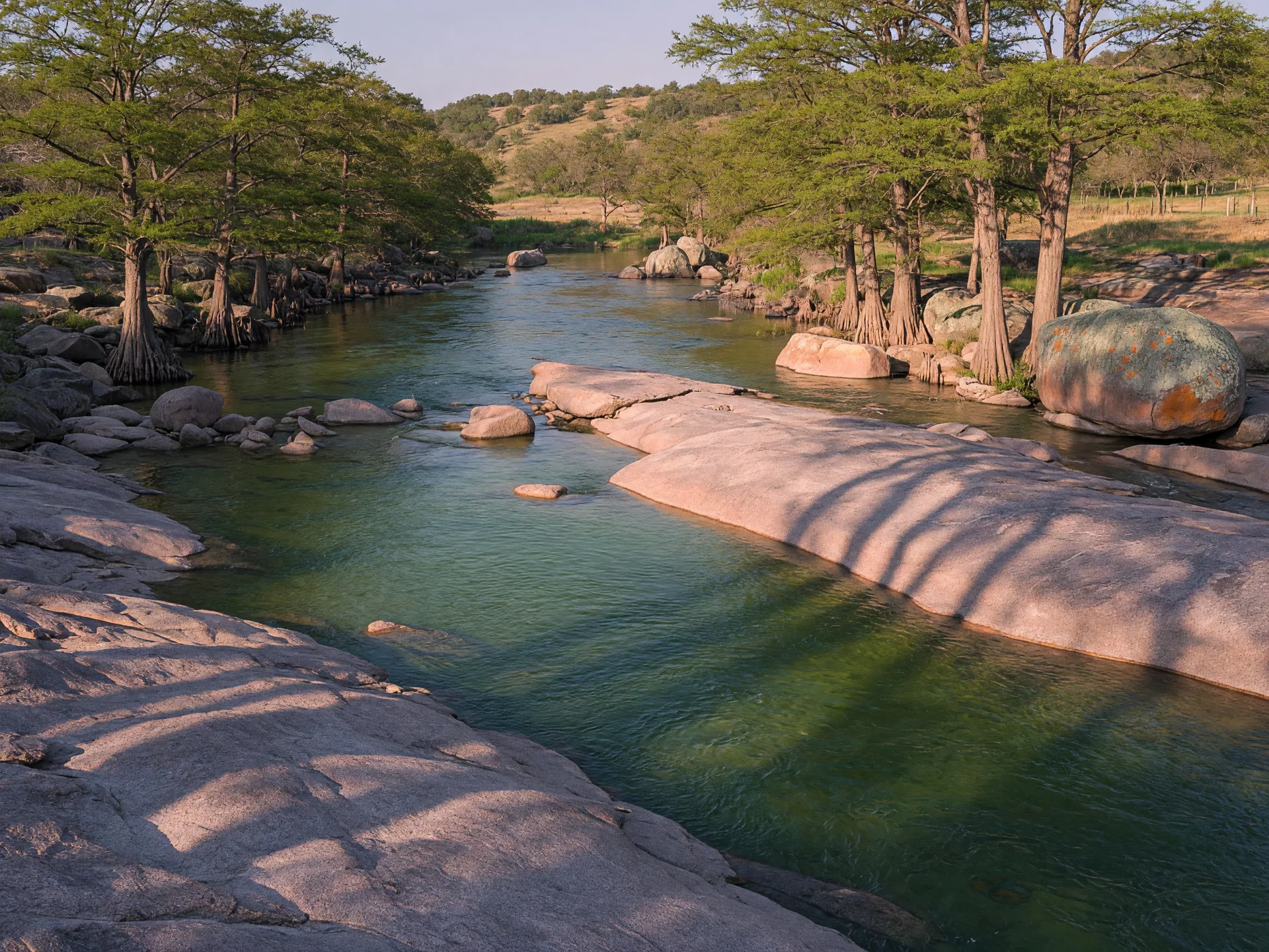 Clear green water of the Llano River flowing over smooth pink-gray granite shelves, with granite boulders and bald cypress trees along the banks under warm late-afternoon light