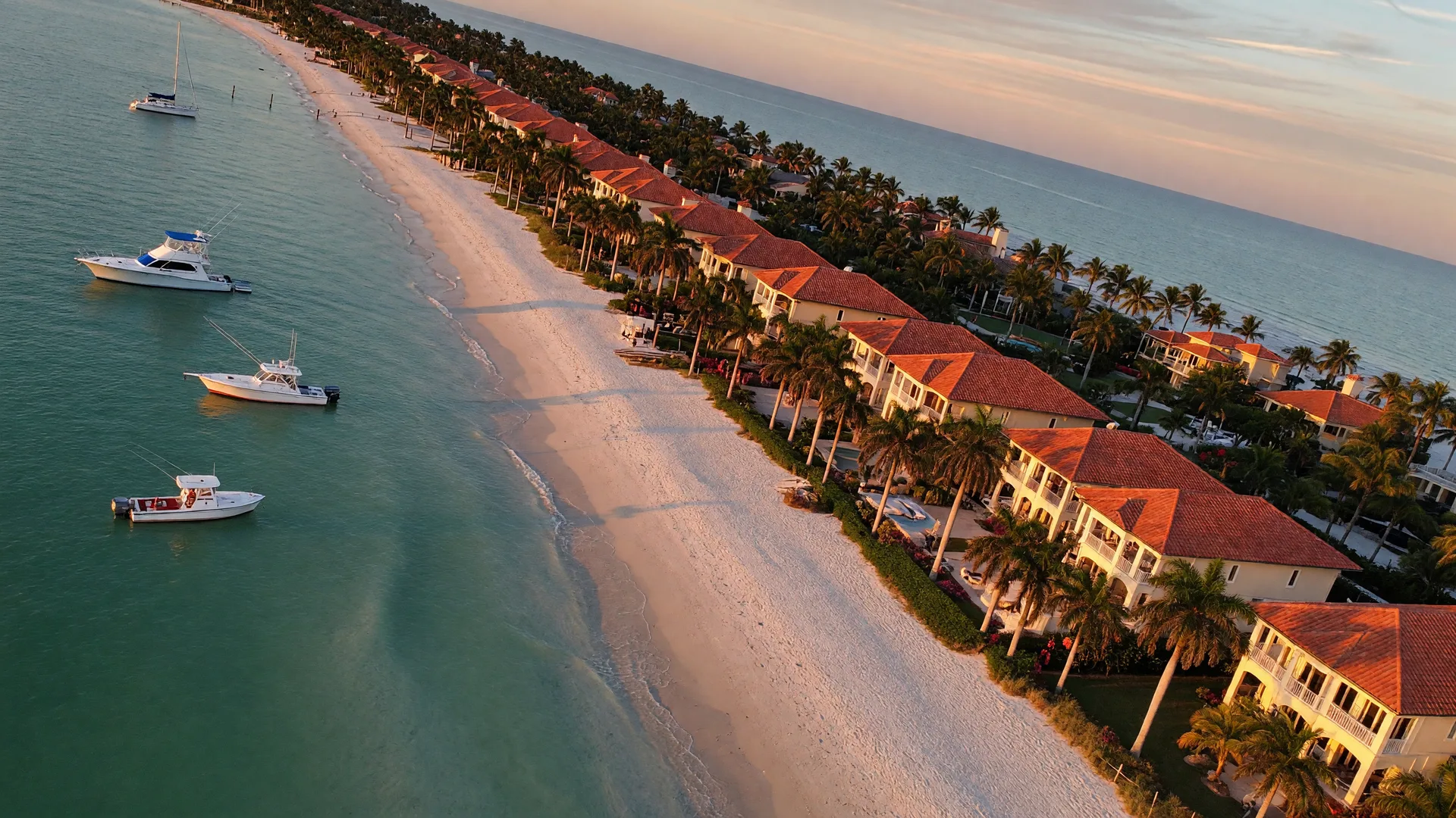 Aerial view of the Naples, Florida coastline at golden hour with Gulf-front Mediterranean homes, white sand beach, royal palms, and calm turquoise water