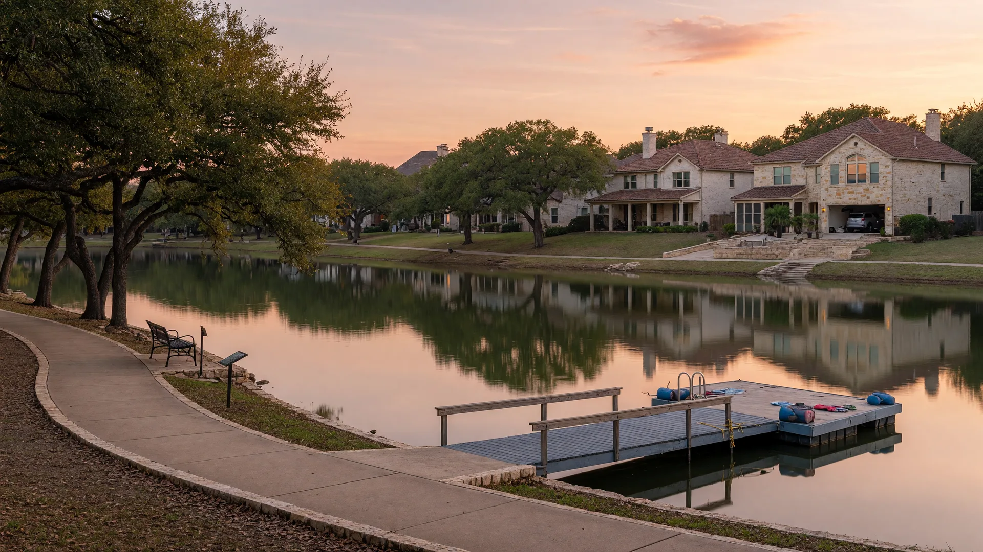 A calm residential lake at golden hour with brick homes on the far bank, a dock, and a walking trail