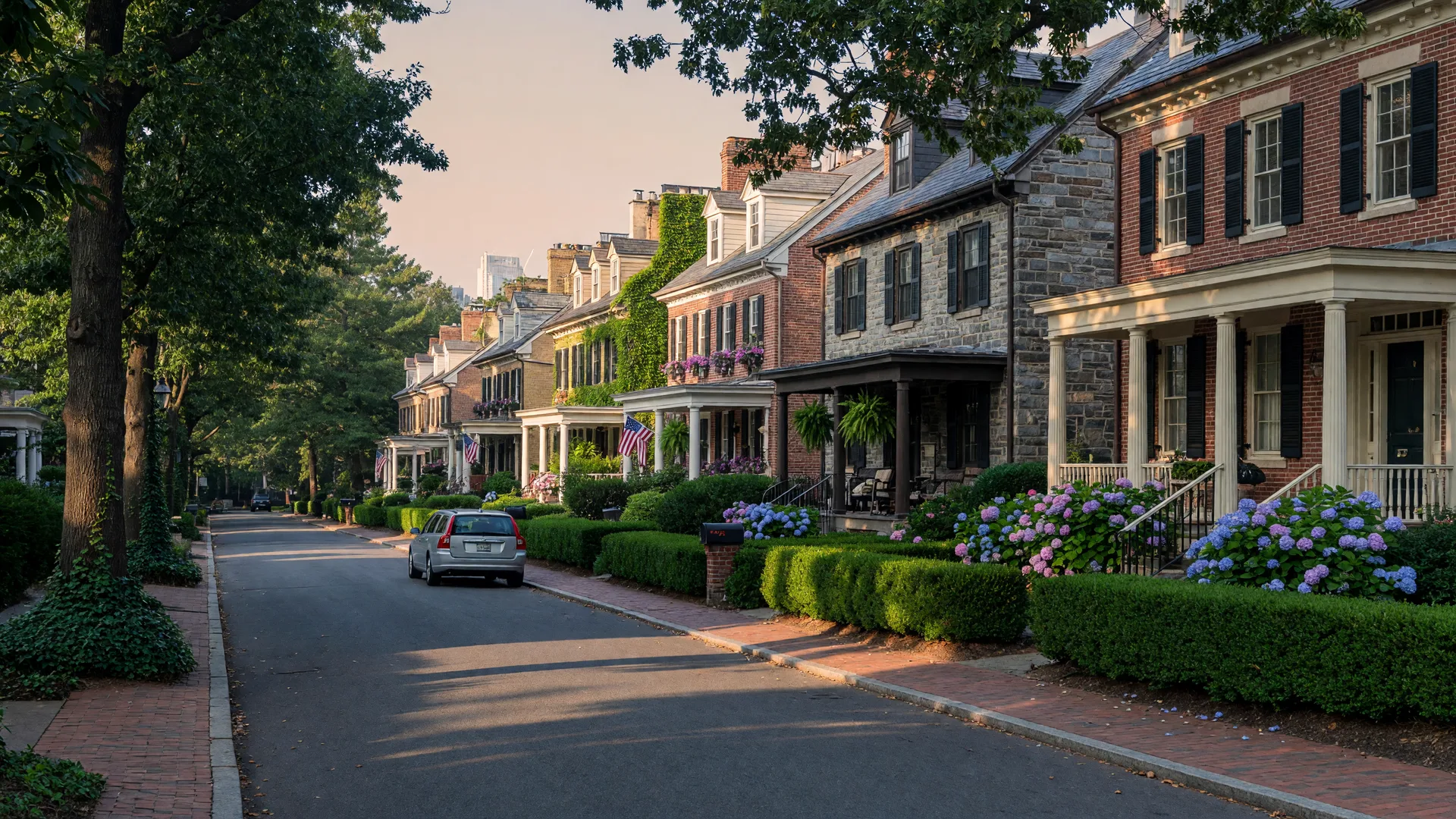 A leafy Chevy Chase street with stately brick and stone colonial homes, hedges and hydrangeas, and mature oaks in late-afternoon light