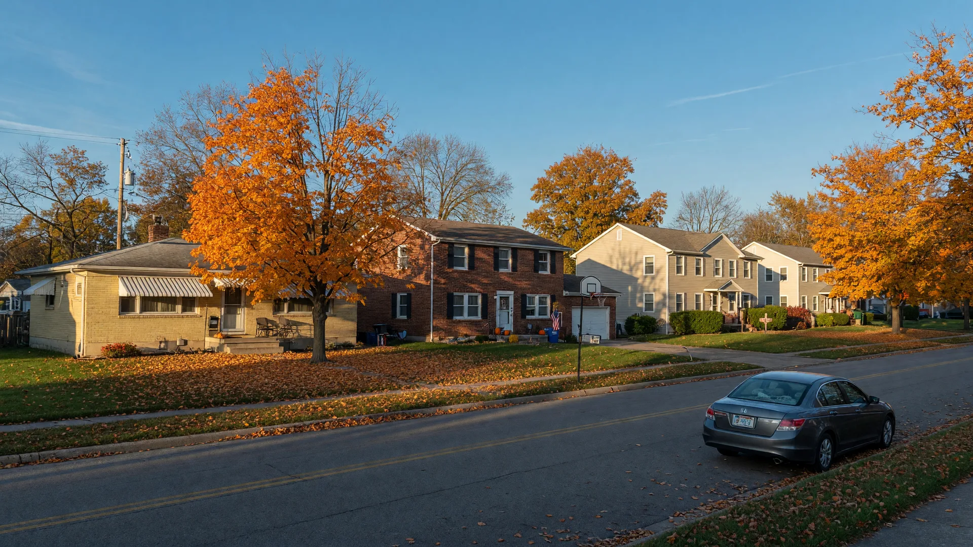 A quiet Fairborn, Ohio residential street on a crisp late-October morning, mid-century brick ranch and two-story homes from the 1950s and 1960s with mature maples and oaks turning orange and gold, newer houses rising on the horizon near Wright-Patterson Air Force Base