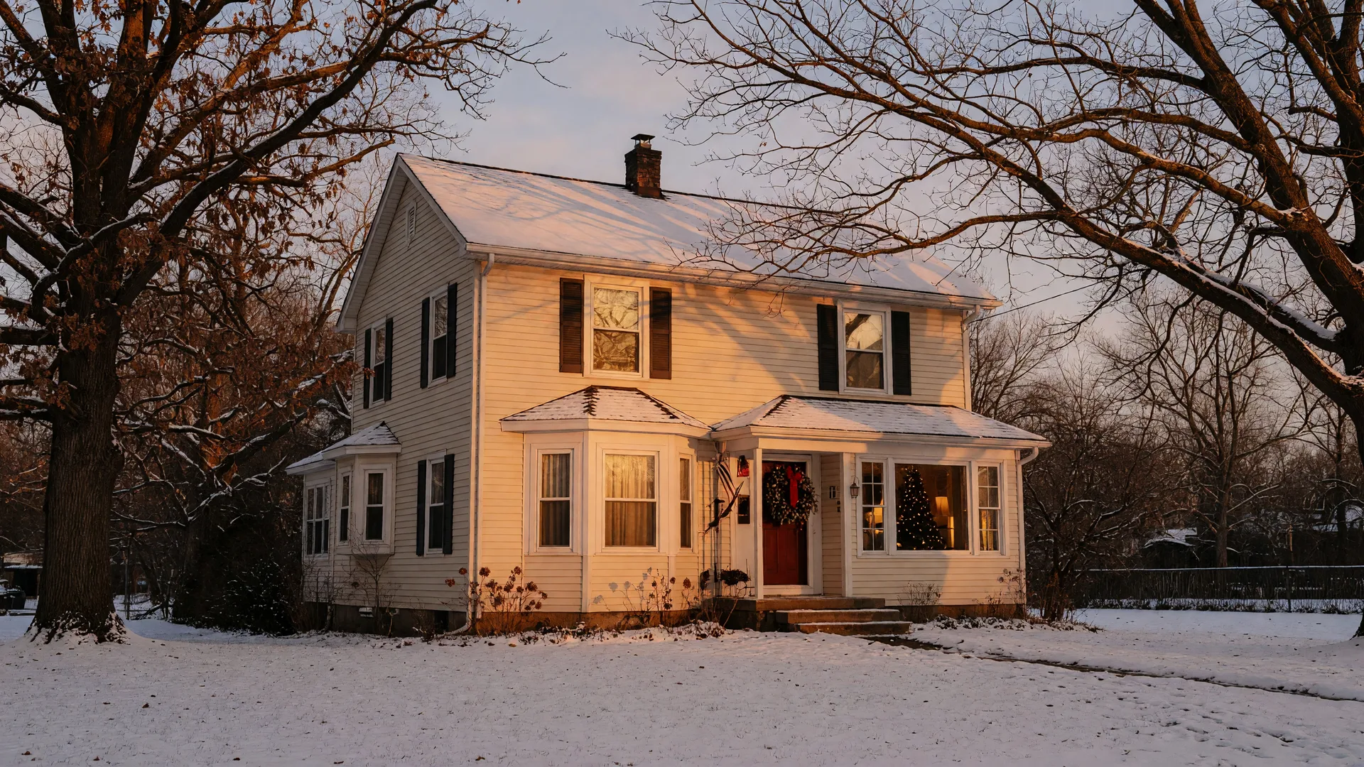 A warm winter photograph of a classic two-story family home in Northwest Indiana with an evergreen wreath on the door, glowing windows, and a light dusting of snow on the lawn