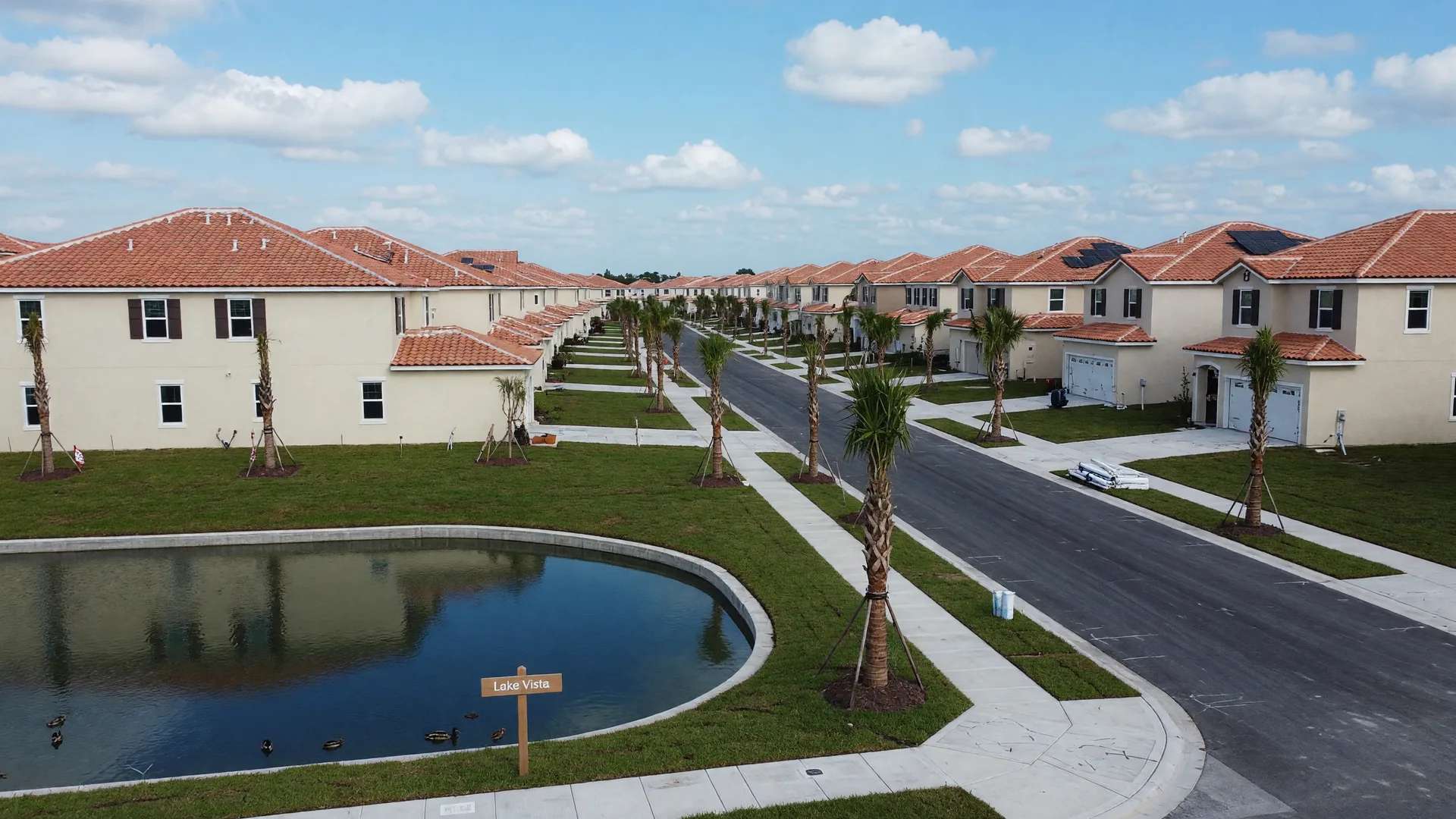 New single-family homes with terracotta roofs and young palm trees lining a freshly paved street beside a lake in a Parrish master-planned community