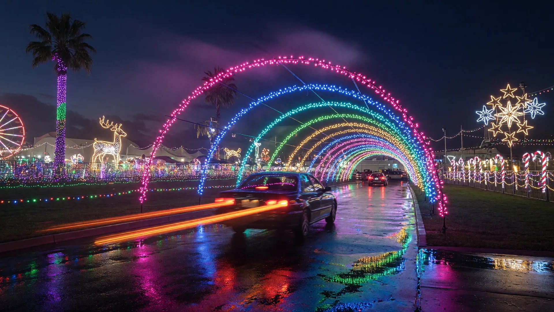 A drive-thru holiday light show at night with a car driving through a tunnel of glowing holiday lights arching over the road under a dark sky