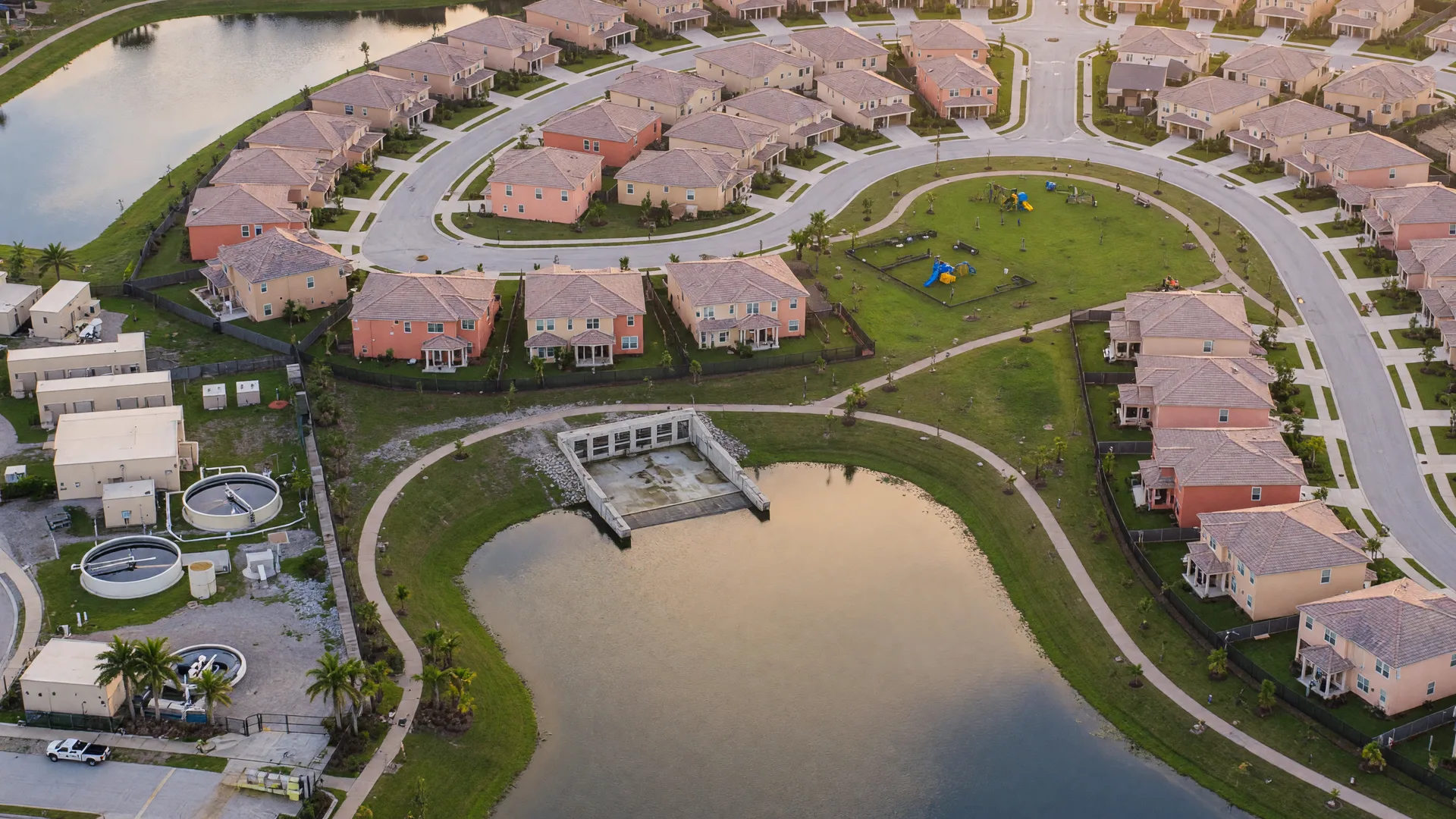 A planned Florida community seen from above at golden hour: new tile-roofed homes along curving streets, a retention lake, and a neighborhood park sharing the same infrastructure.