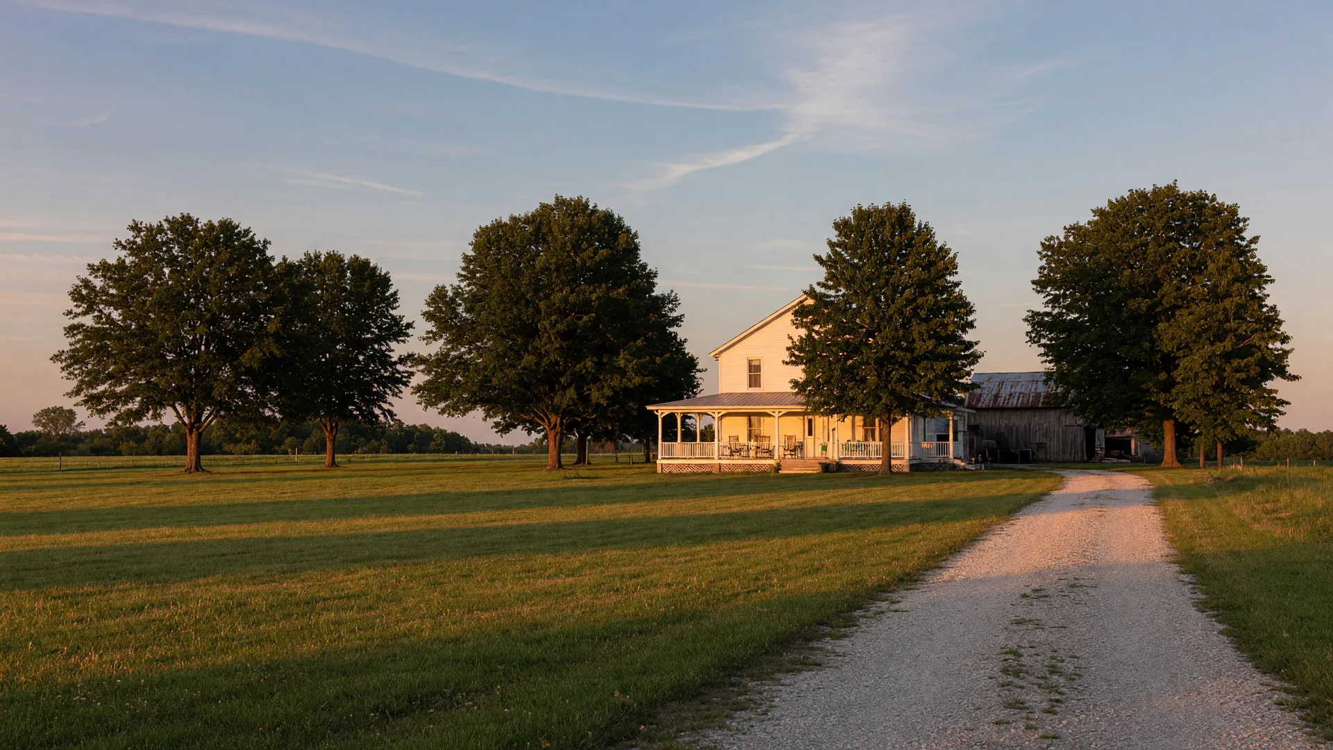 A white Kentucky farmhouse with a wraparound porch on open green acreage at golden hour