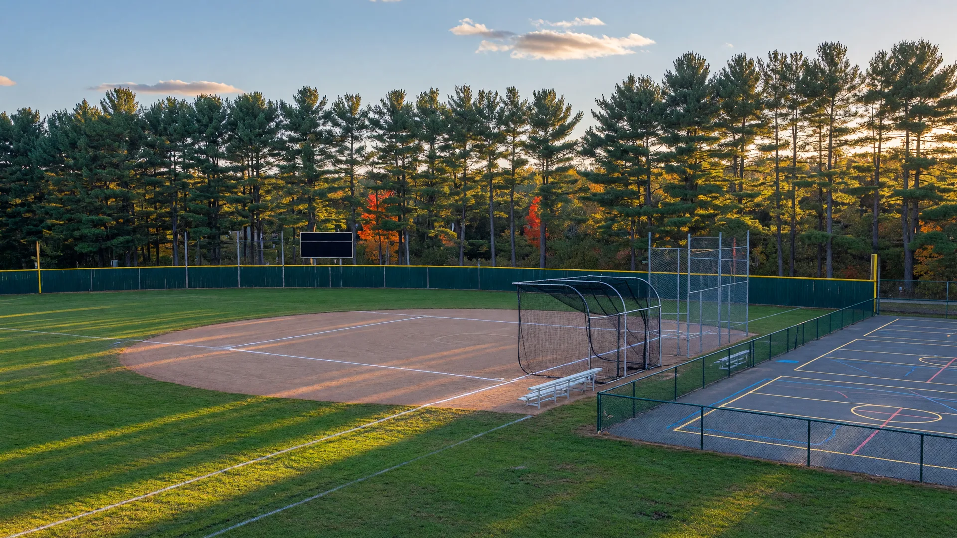 Community ballpark with a batting cage and multi-use sport courts beside pines on a crisp September afternoon