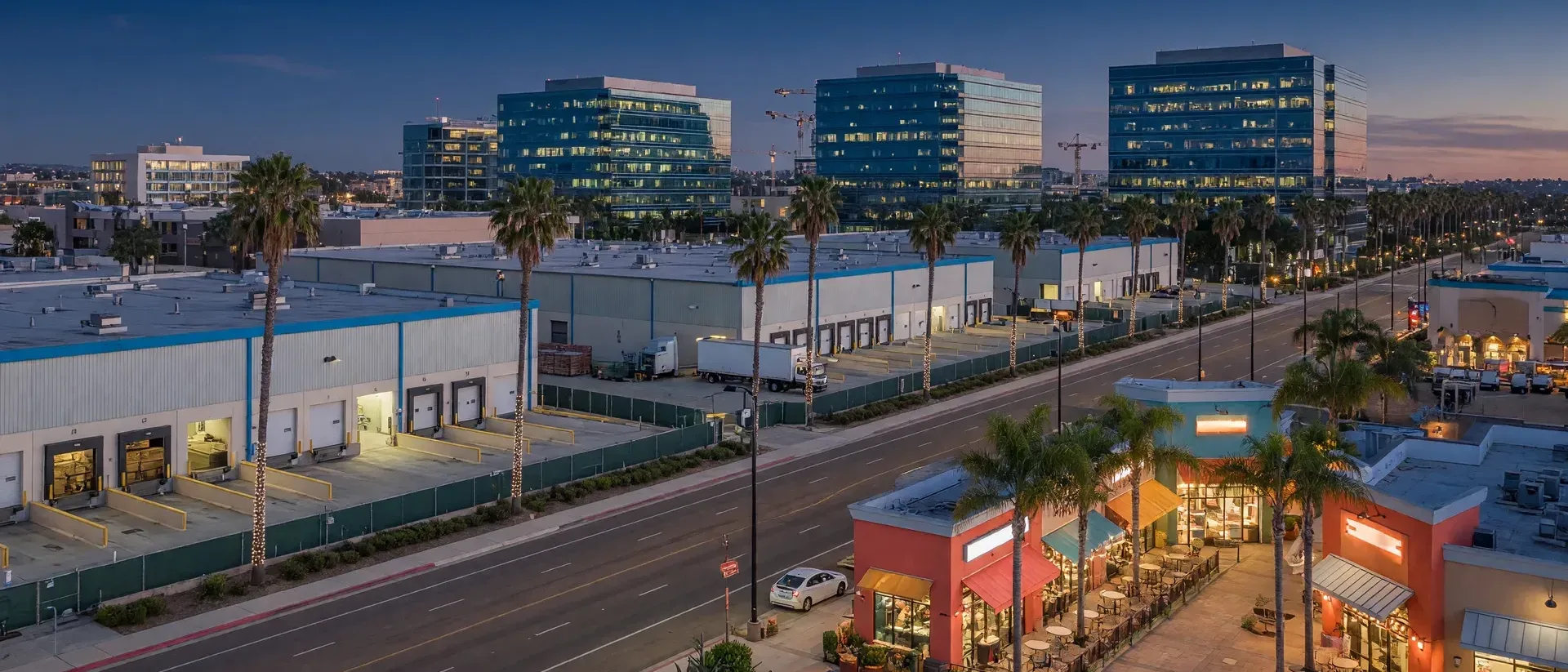 San Diego commercial district at dusk with glass office towers, distribution warehouses, and a glowing retail plaza along a palm-lined boulevard