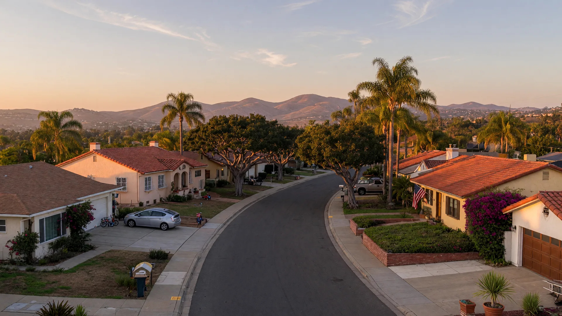 Aerial-style view of a sunny North County San Diego residential neighborhood at golden hour, ranch-style and Spanish tile-roofed single-family homes along a tree-lined street with coastal hills in the distance