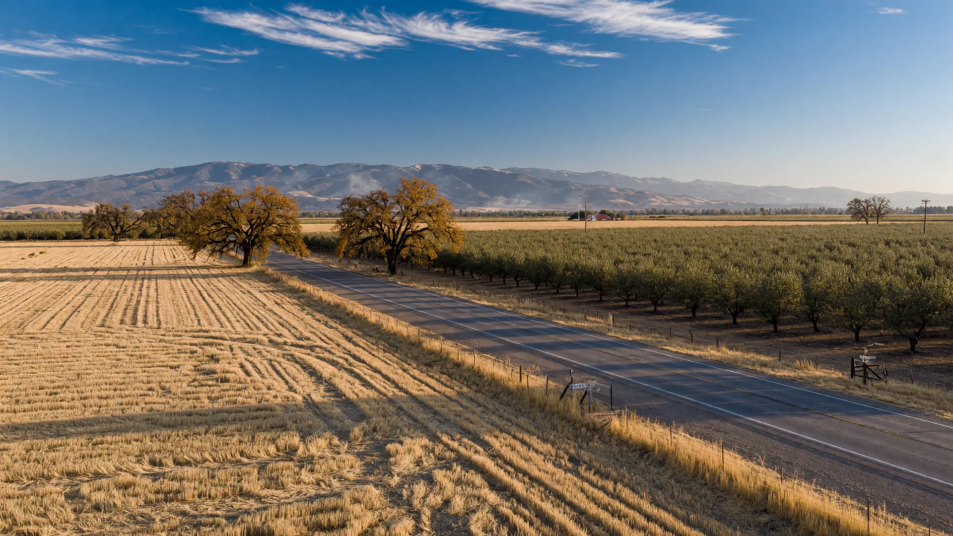 Golden harvested fields, almond orchards, and valley oaks near Merced in the Central Valley on a sunny mid-October morning, with the hazy Sierra Nevada foothills on the horizon under a clear blue sky