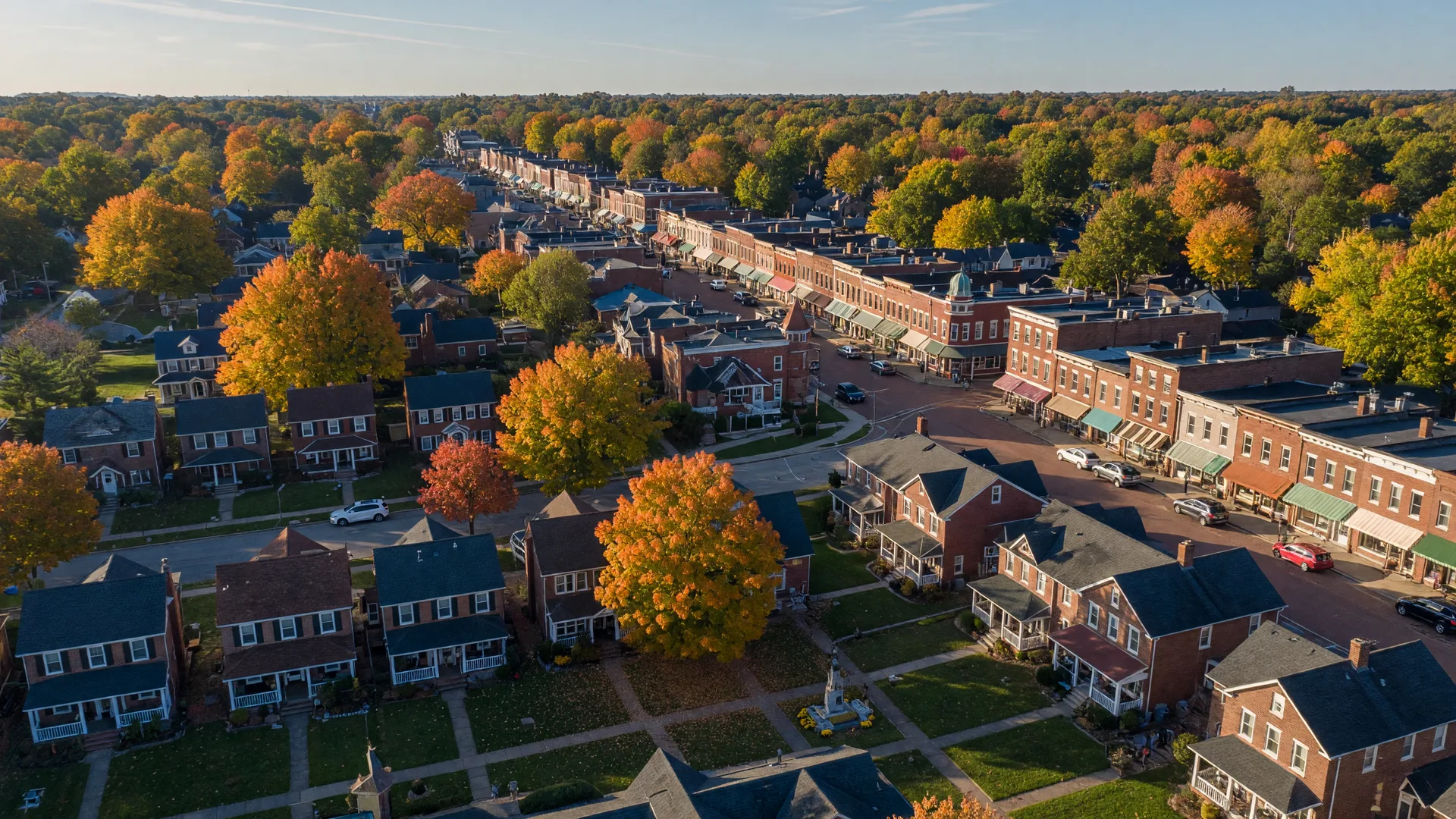 A wide aerial view of Centerville, Ohio in early autumn, looking down over a tree-canopied residential suburb of brick colonials and craftsman homes with maples turning orange and gold, a historic downtown main street with preserved brick storefronts in the middle distance, in warm late-afternoon light
