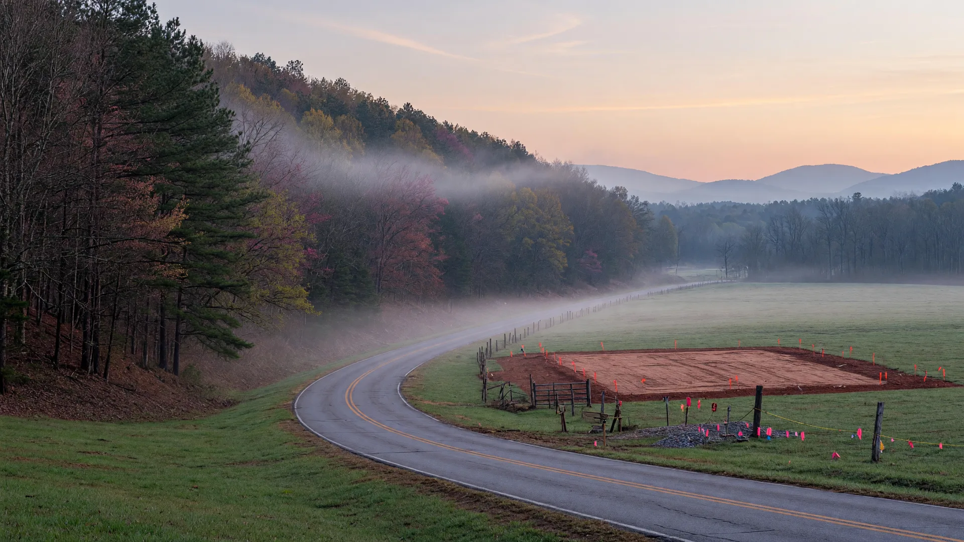 Rural North Georgia county road at dawn with timbered hillside on one side and a freshly graded, stake-and-flag-marked homesite on the other