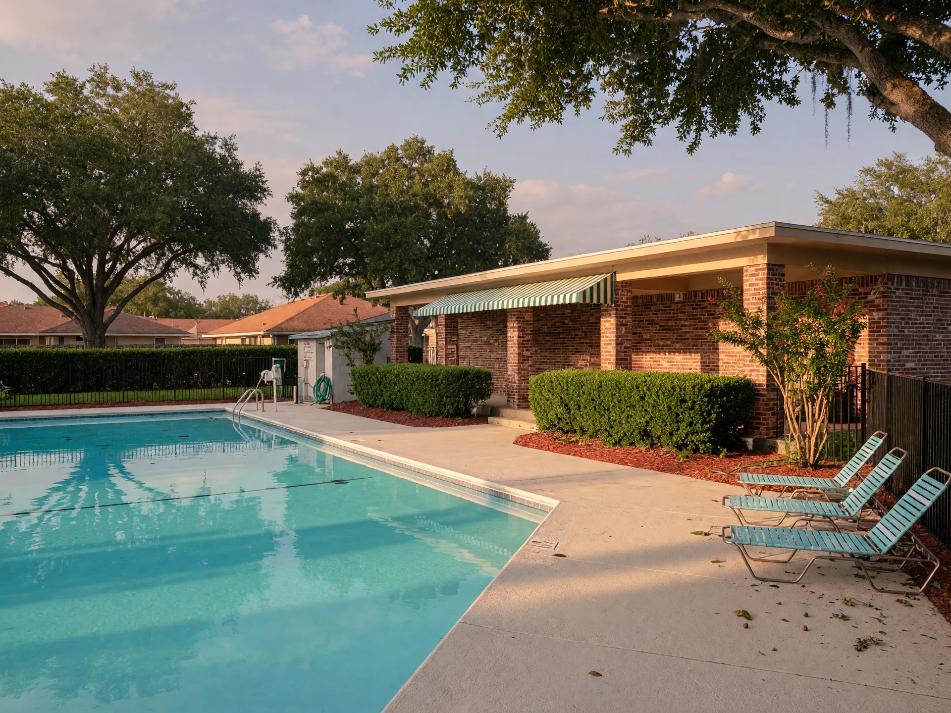 A community pool and modest clubhouse in a residential HOA neighborhood with mature oak trees and lounge chairs