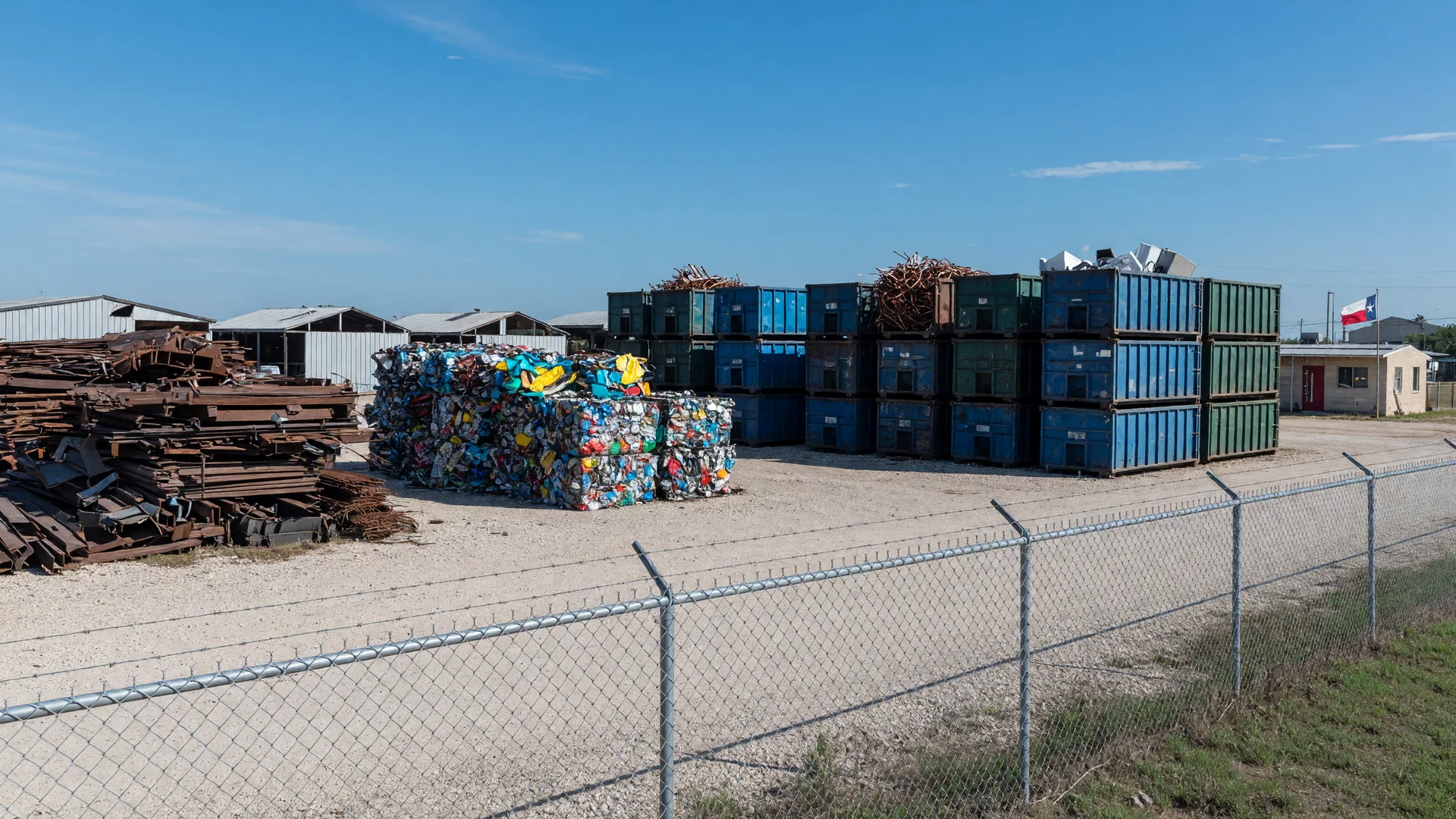 Tidy scrap metal recycling yard in a small Texas town with sorted ferrous and aluminum piles and steel roll-off containers under a clear blue sky