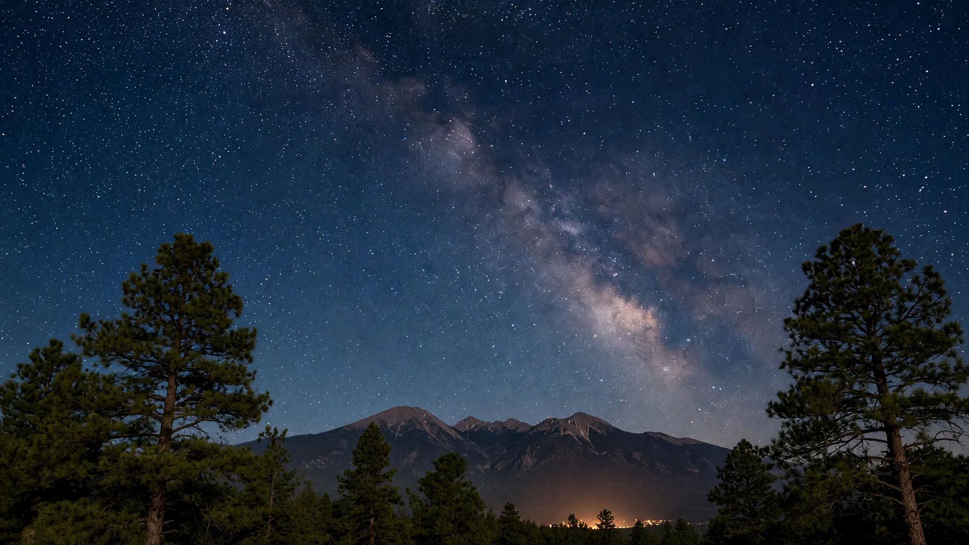 The Milky Way arcing over a dark Flagstaff night sky above ponderosa pines and the San Francisco Peaks