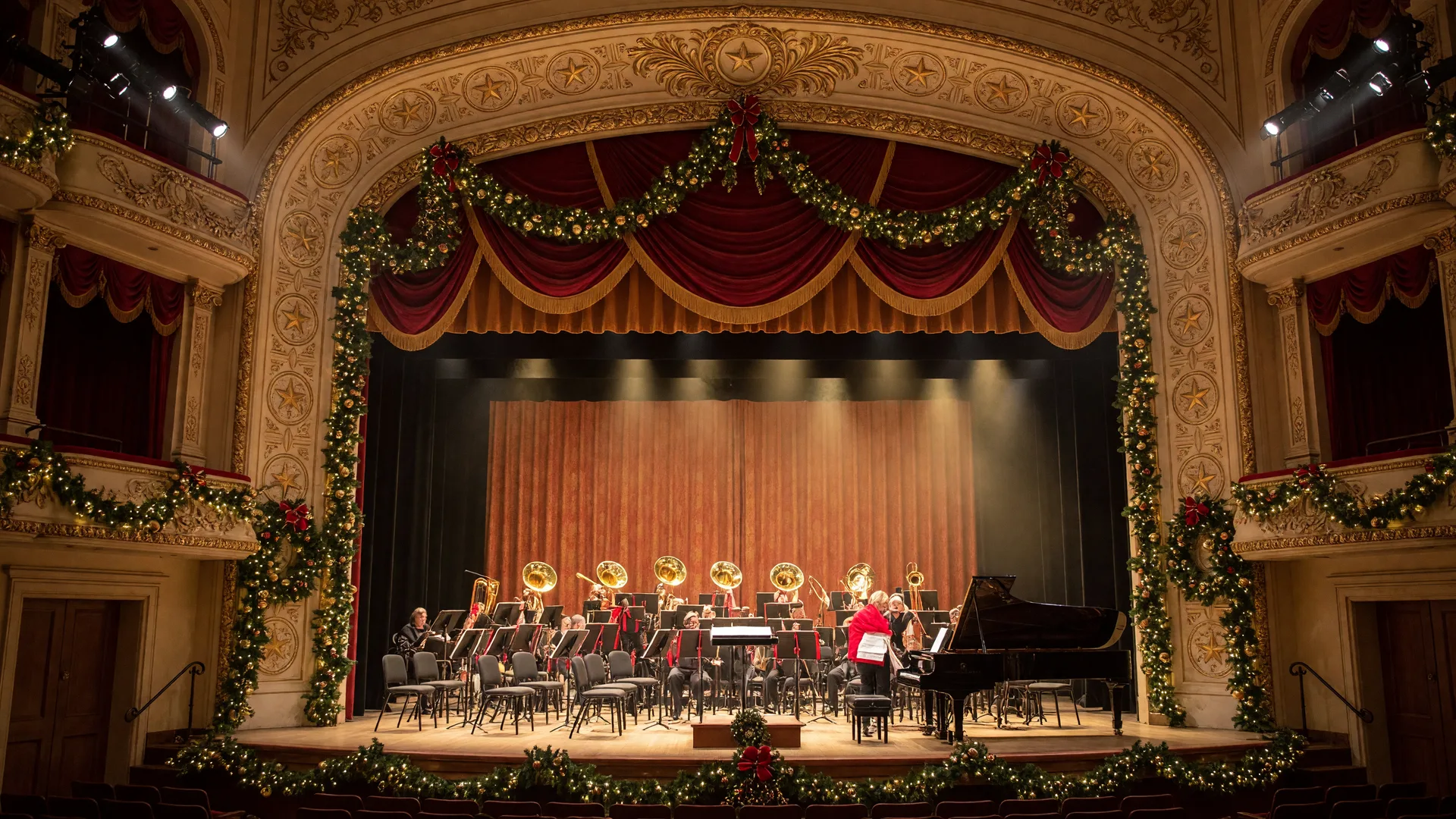 A historic Texas auditorium stage dressed for a Christmas pops concert with a full orchestra, grand piano, evergreen garlands, and warm string lights