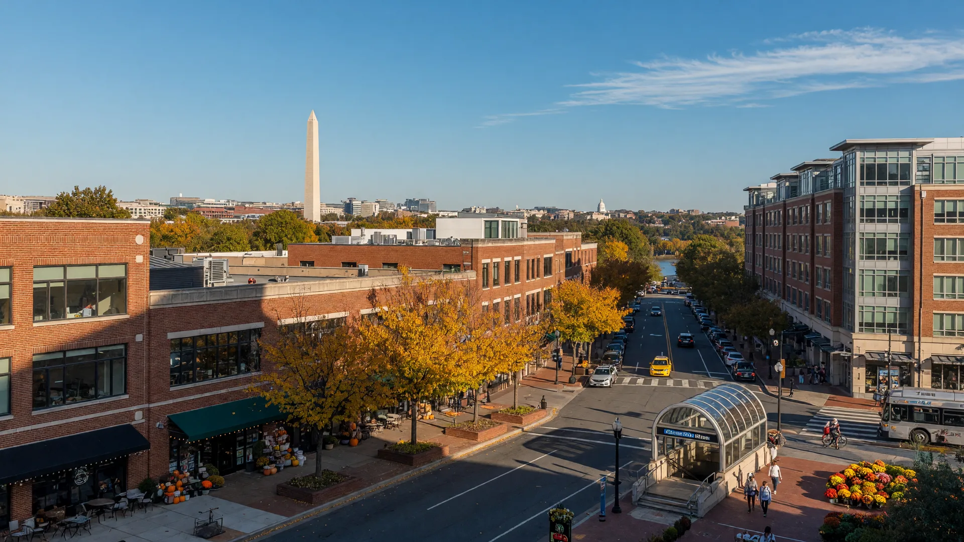 Wilson Boulevard in Arlington, Virginia's Clarendon neighborhood on a golden October afternoon, brick and glass urban village storefronts with cafes and shops, fall trees, a Metro station entrance, and the Washington Monument and DC skyline visible in the distance