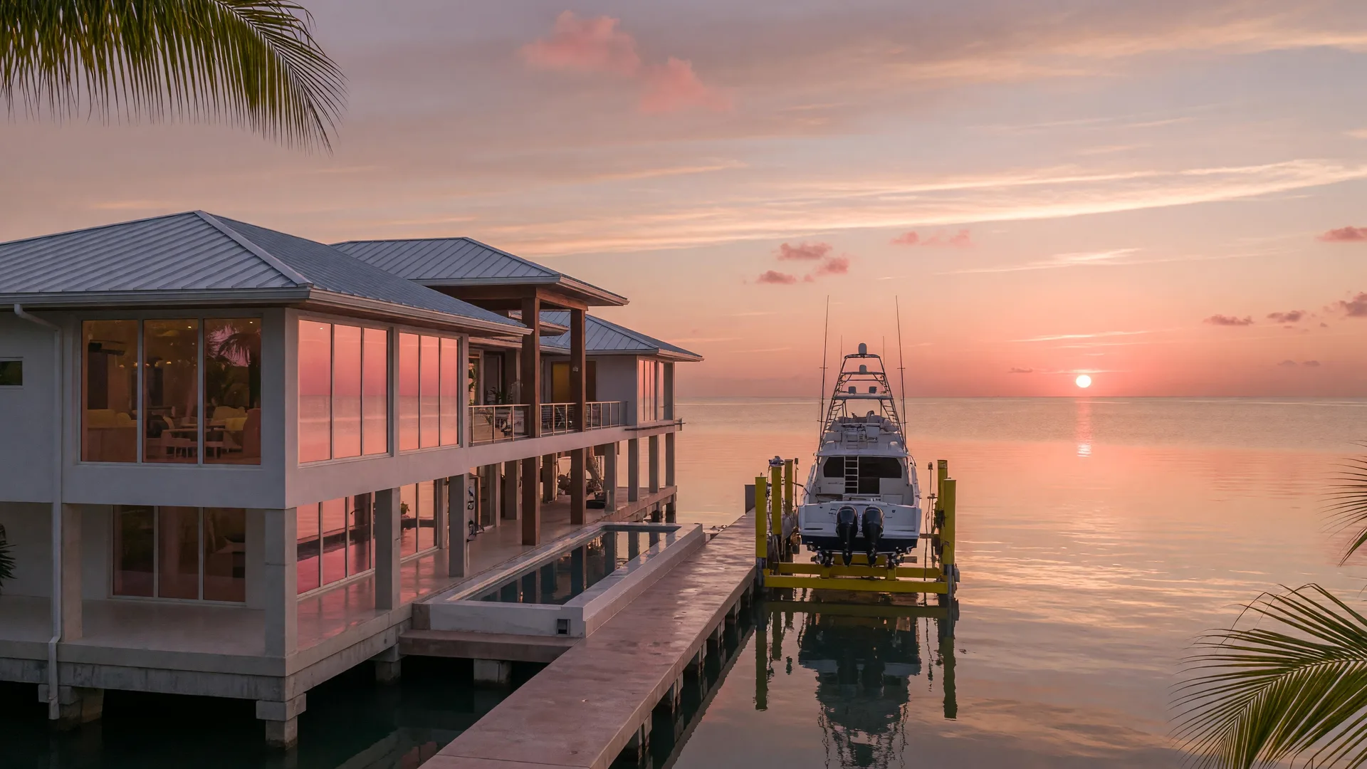 A luxury east-facing oceanfront estate in the Florida Keys at sunrise, an elevated contemporary home with a standing-seam metal roof and floor-to-ceiling impact glass catching pink-gold light, a private concrete dock with a boat lift and sportfishing yacht, open Atlantic water with the sun low on the horizon