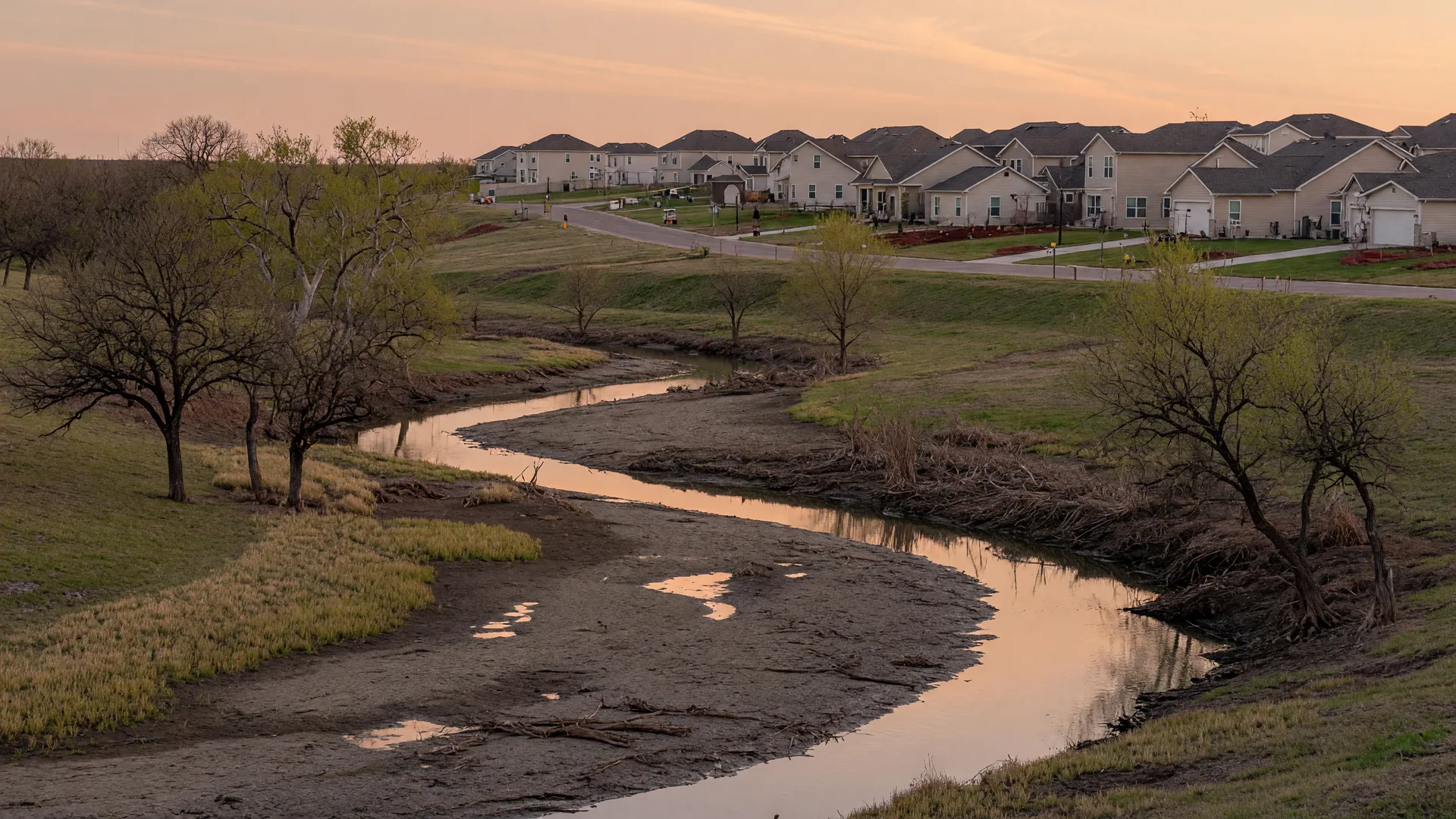 A slow-moving creek winding through a North Texas floodplain at golden hour, with new homes on higher ground in the distance