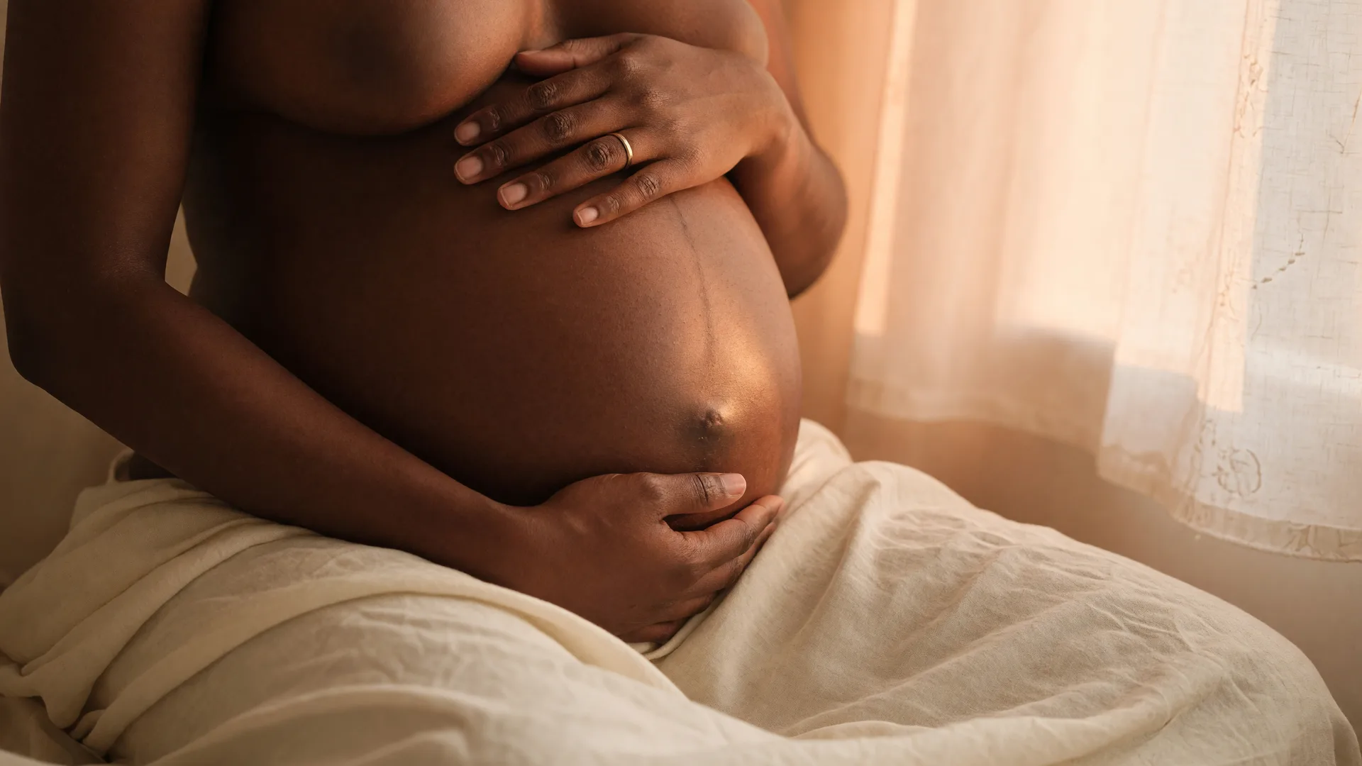 A Black mother's hands gently cradling her pregnant belly in warm golden light