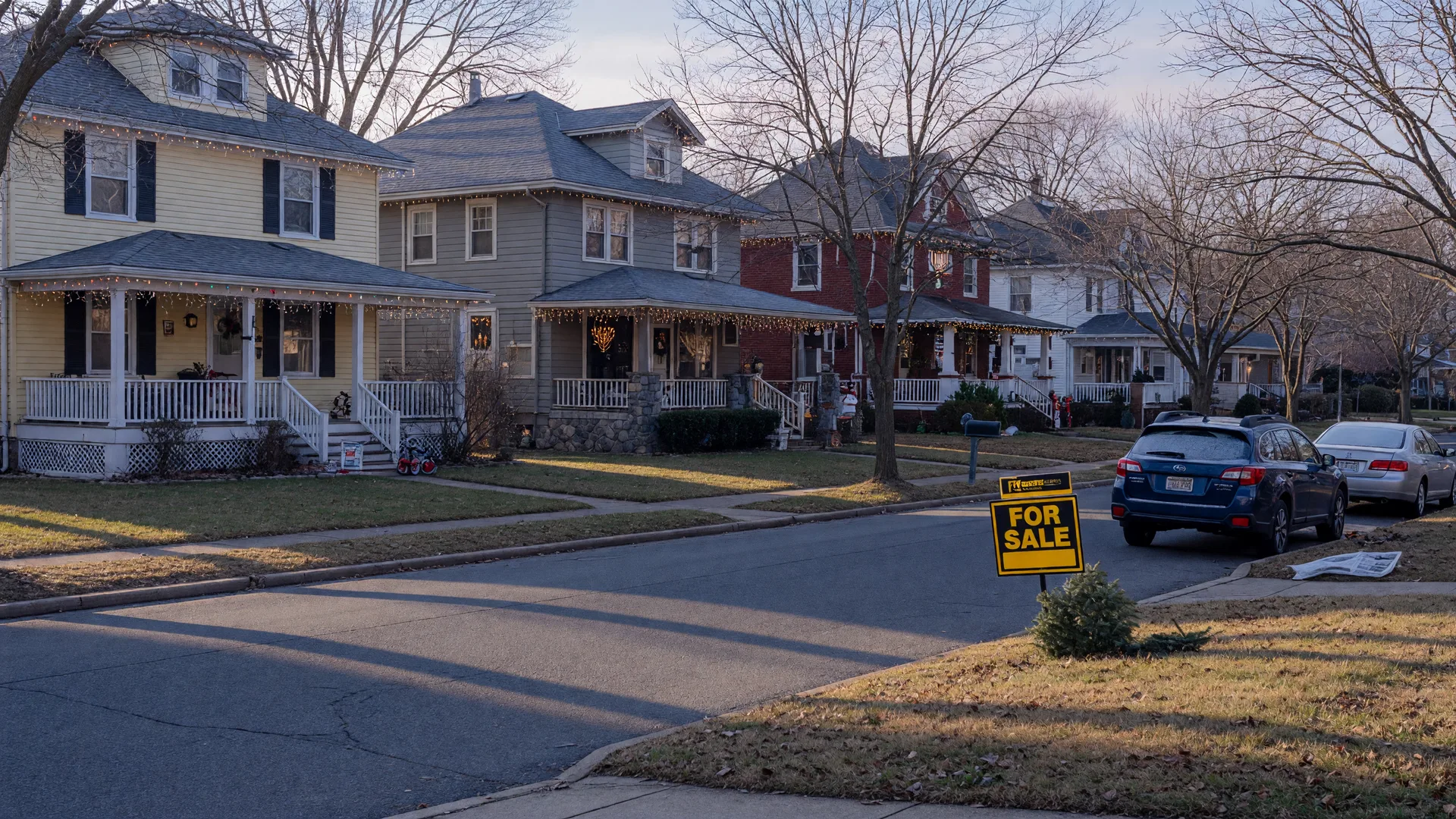 A quiet New Jersey suburban street on a crisp December afternoon with colonial homes wearing warm holiday porch lights, a real estate for-sale sign on a front lawn, and bare trees casting long winter shadows