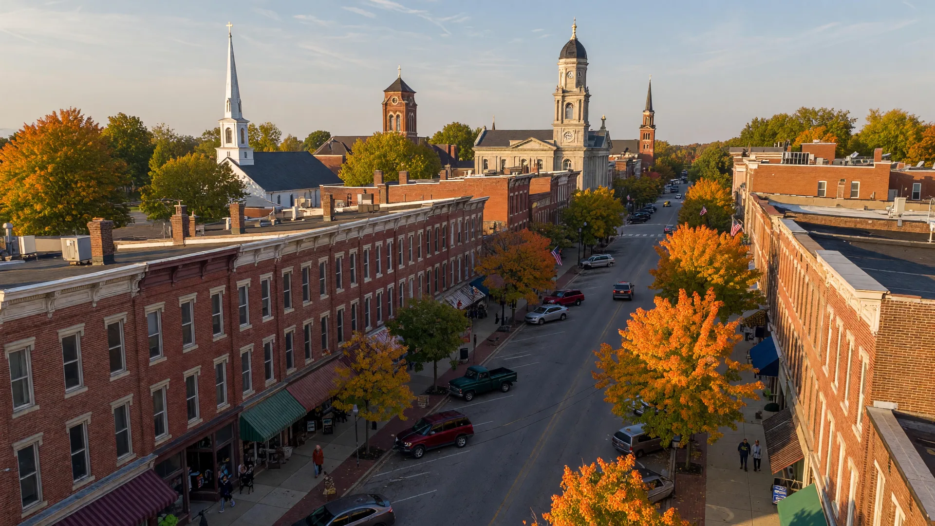 A wide aerial view of historic downtown Lebanon, Ohio in early autumn, looking down the tree-lined Broadway main street of preserved 19th-century brick storefronts, church steeples and the courthouse rising above the roofline, and maples turning orange and gold in warm late-afternoon light