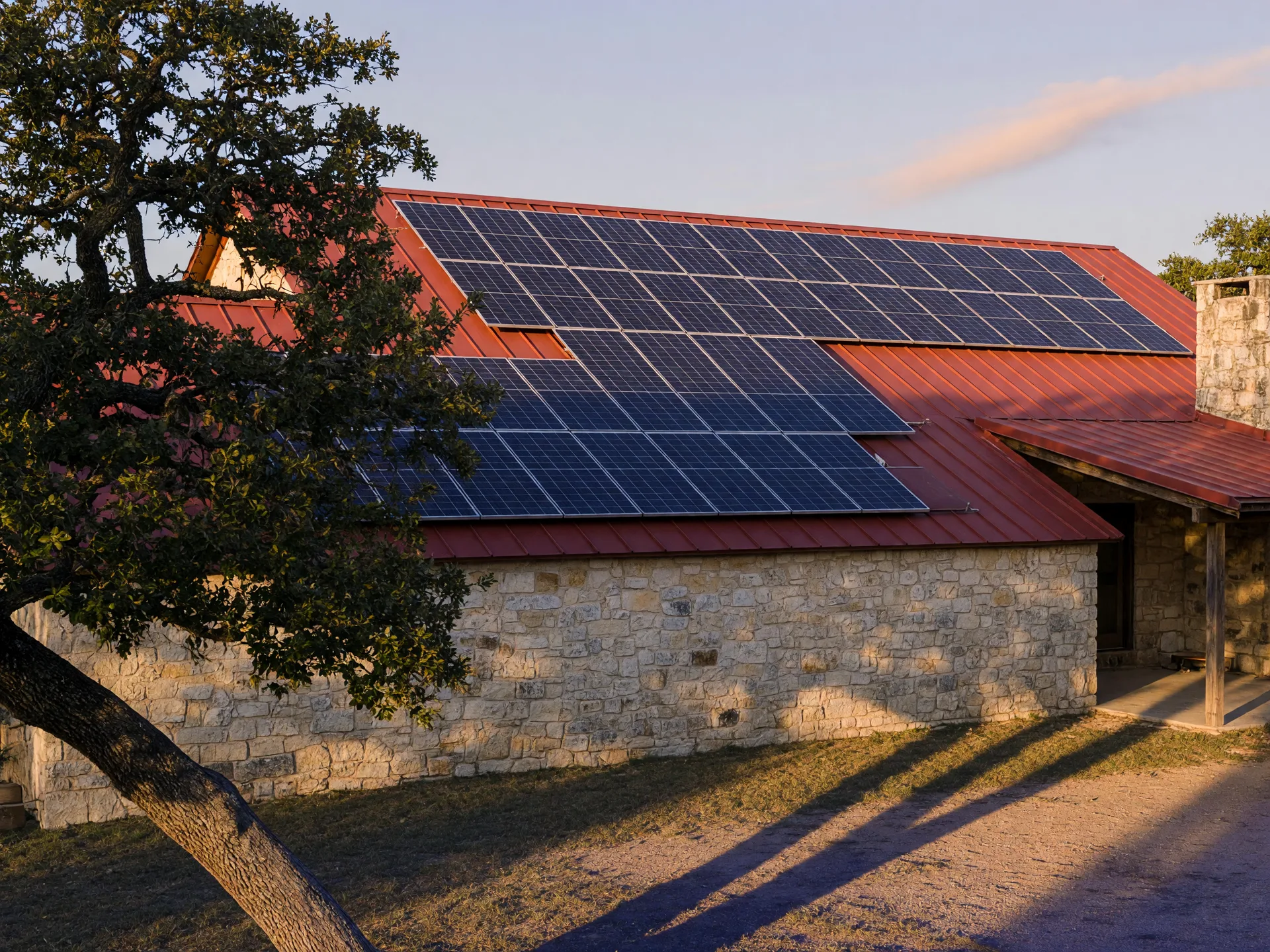 Solar panels installed on the standing-seam metal roof of a Texas Hill Country limestone farmhouse at golden hour, with a mature oak tree in the left foreground