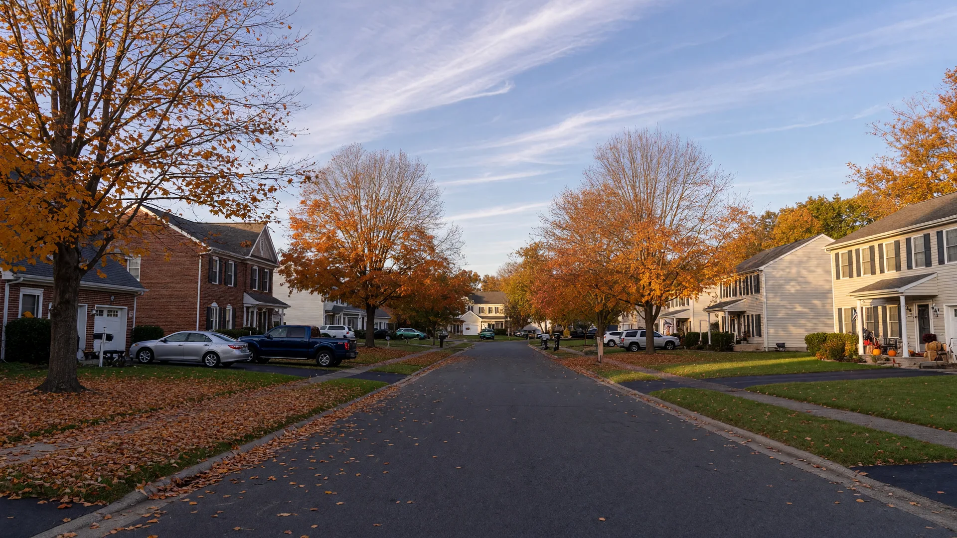 A tree-lined suburban street in South Jersey in mid-October with brick colonial homes under amber and orange fall foliage