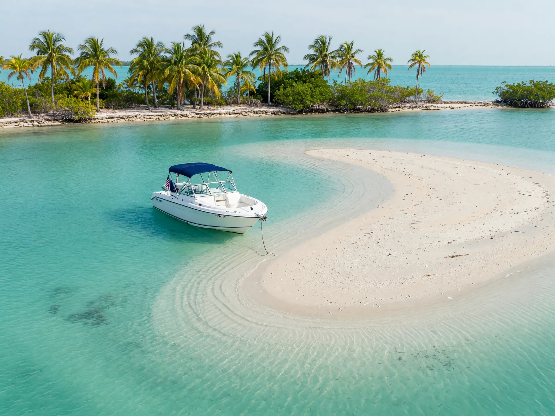 A boat anchored in clear turquoise shallows beside a white sandbar off a palm-lined island in the Upper Florida Keys, calm midday water with no people