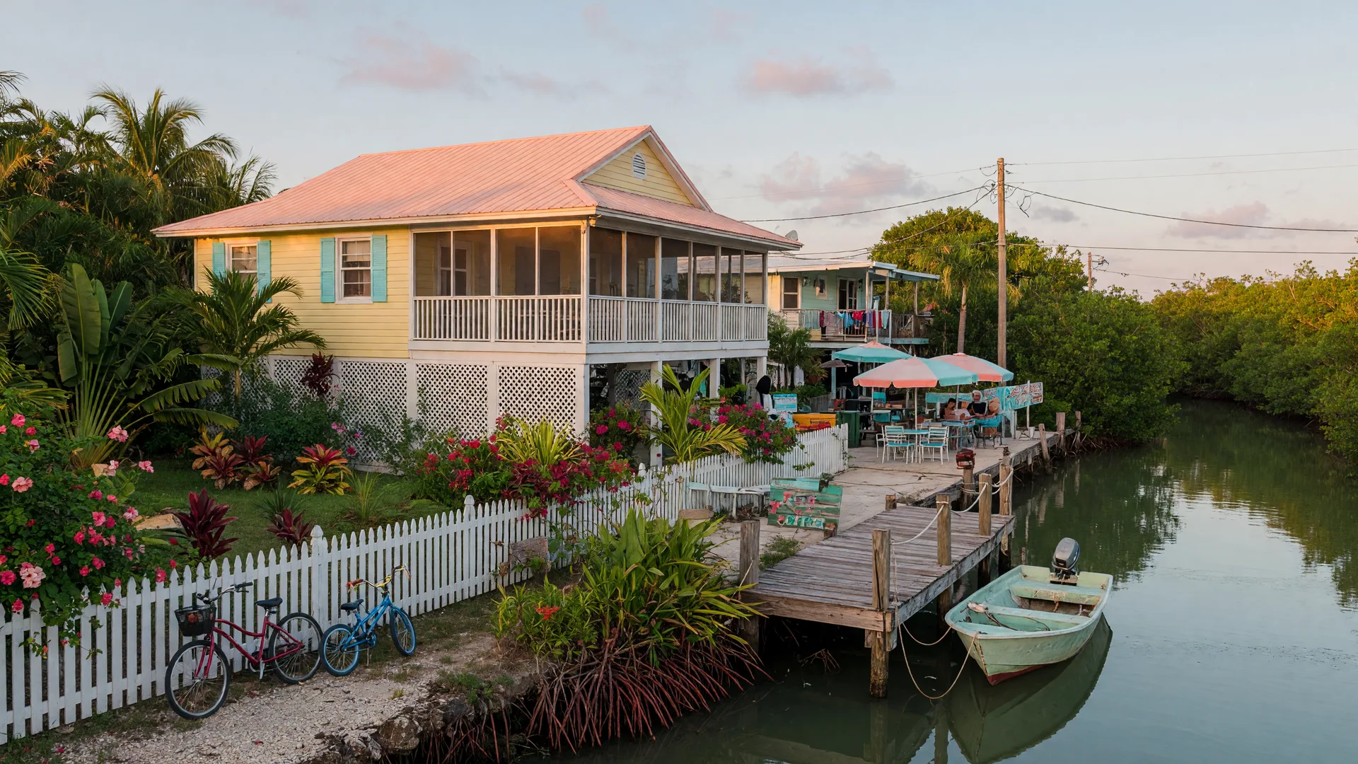 A golden-hour residential street in the Upper Florida Keys, an elevated metal-roofed Keys home with a lush tropical yard and a white picket fence, bicycles by the front walk, a small cafe patio with umbrellas in the near distance, and a calm mangrove-lined canal with a small skiff behind