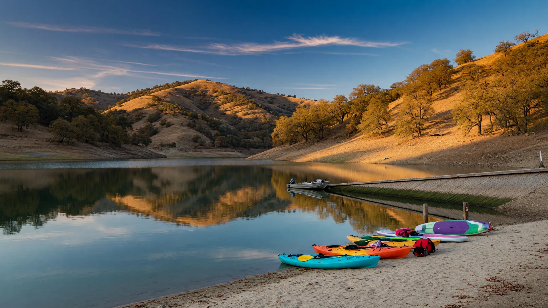 A calm Sierra-foothill reservoir in the Central Valley on a clear late-October afternoon, golden oak-covered hillsides, a sandy swim beach with colorful kayaks and paddleboards pulled up on the sand, glassy water reflecting the amber hills, and a wooden boat launch ramp