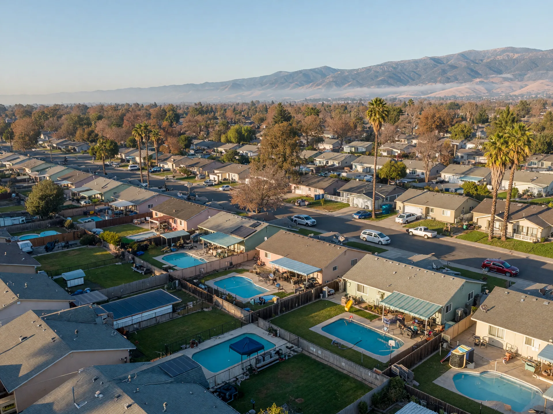 An elevated view of a Merced County residential neighborhood on a clear October morning, rows of ranch and tract homes, several backyards with in-ground swimming pools and covered patios, and the hazy Sierra Nevada foothills on the horizon