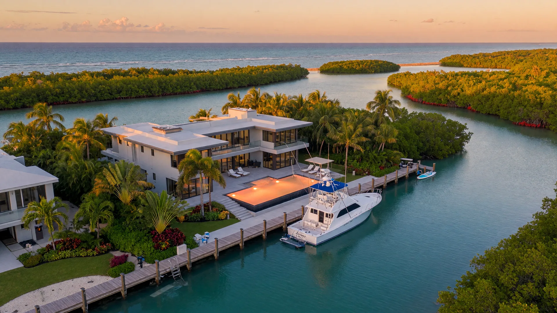 An aerial view of a luxury deep-water canal estate in Islamorada at golden hour with a contemporary waterfront home, private dock, sportfishing yacht, infinity-edge pool, and turquoise canal water