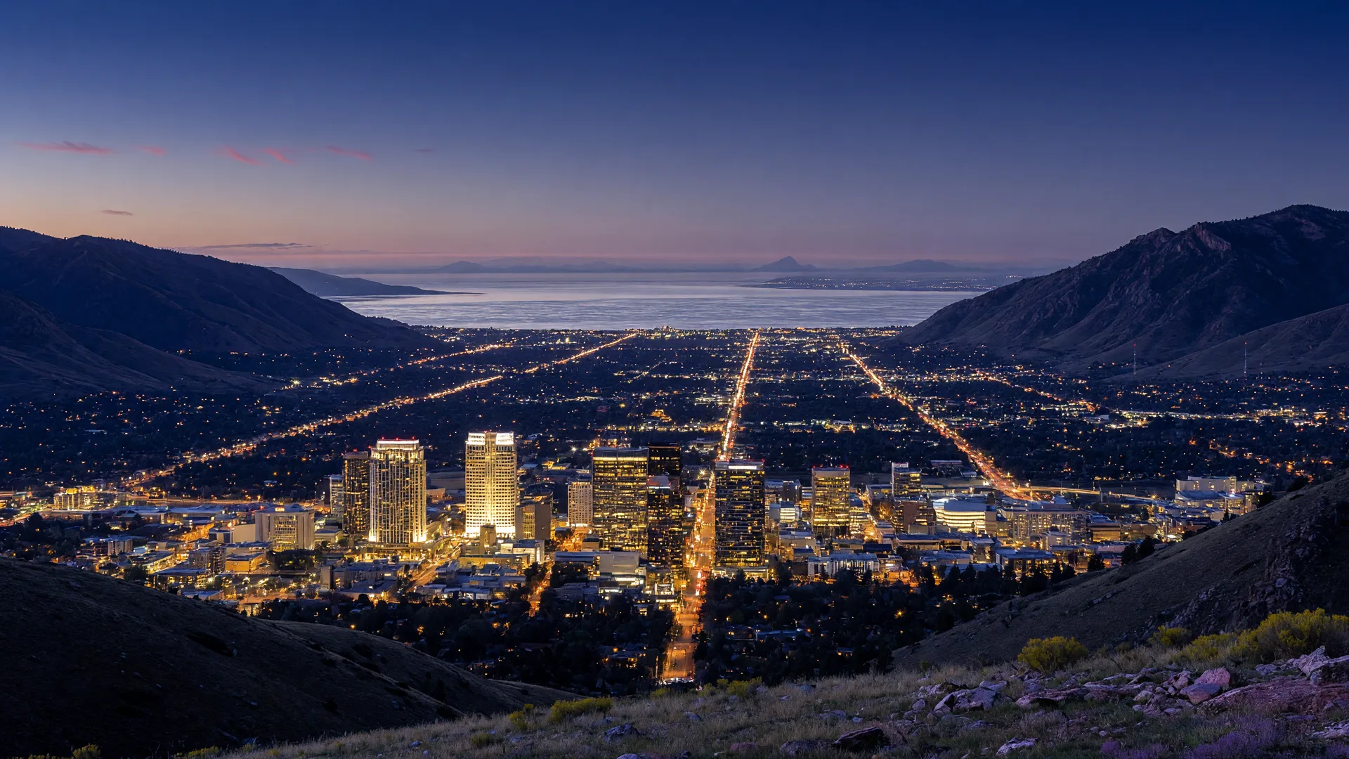 Salt Lake City's skyline at blue hour seen from the Wasatch foothills, downtown towers glowing against an indigo sky with the Great Salt Lake on the horizon.