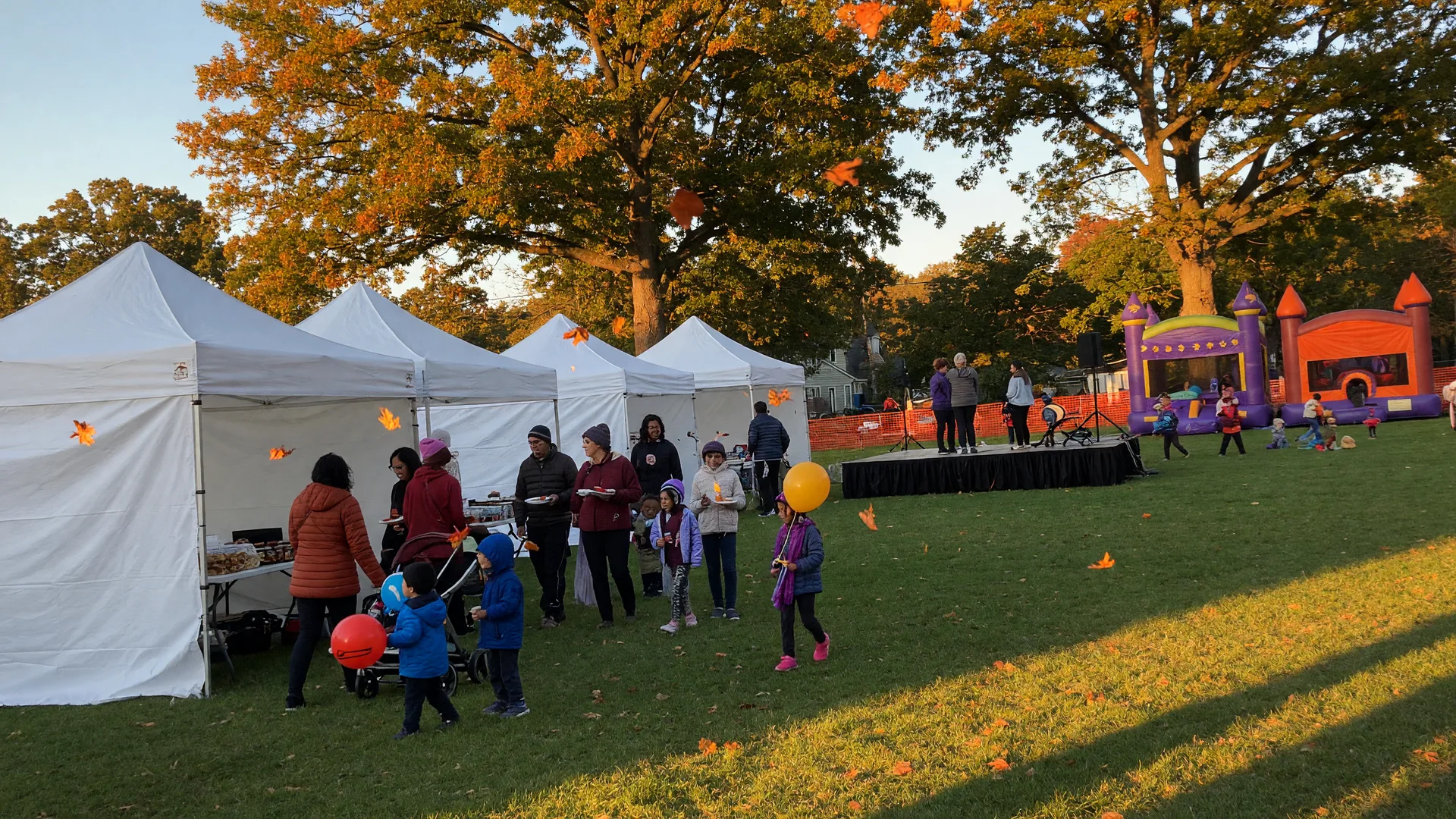 A township fall festival on a crisp early-October afternoon with white vendor tents and a small stage on a green lawn, families strolling with kids holding balloons, bounce houses in the distance, and mature oaks in gold and orange overhead in warm golden-hour light