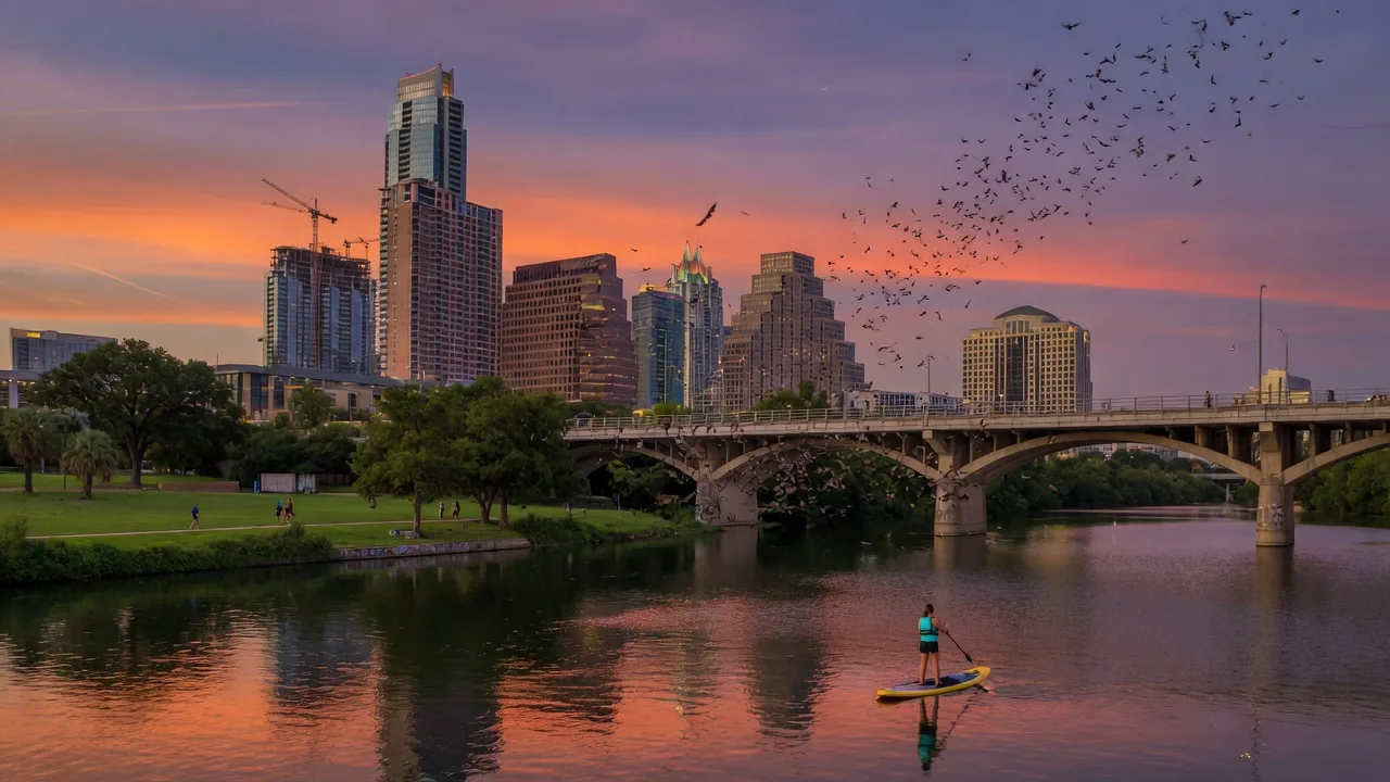 Austin Texas skyline at sunset — Lady Bird Lake, Congress Avenue Bridge, and downtown towers reflected in the water under warm purple and orange skies