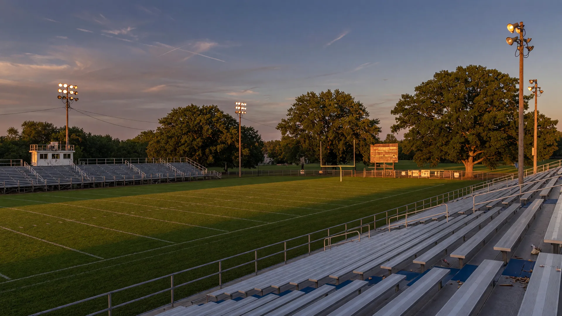 A late-summer evening at a rural Clinton County high school football stadium at golden hour with freshly painted goalposts, oak trees, and stadium lights beginning to glow against a deepening blue sky