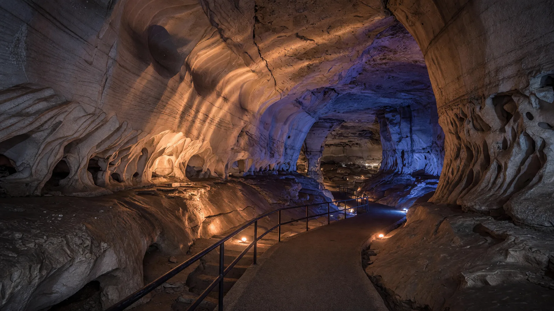 Warmly lit cathedral-like limestone chamber inside Longhorn Cavern with smooth river-carved walls and a descending paved walkway with a metal handrail