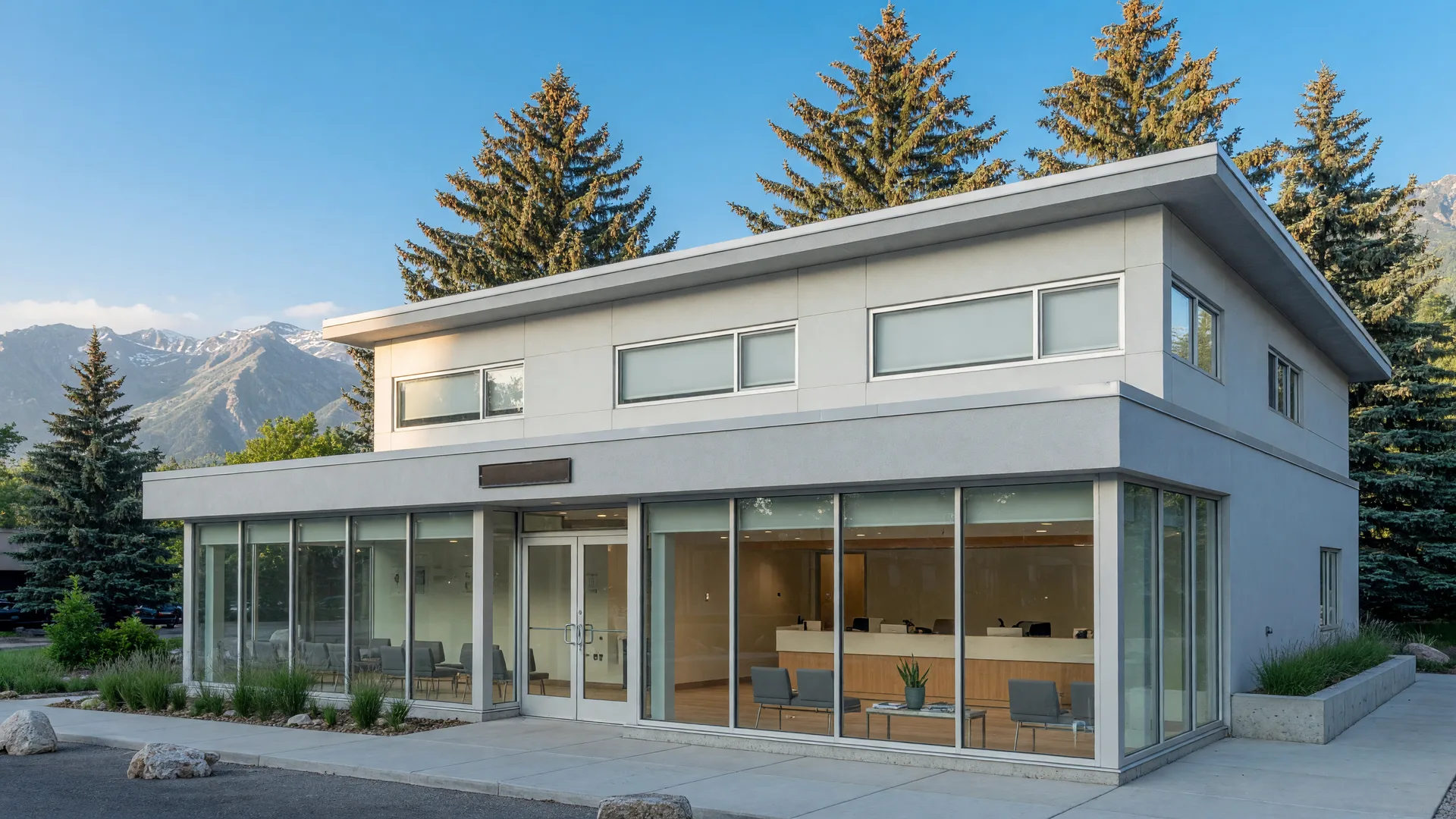 A bright, modern medical office building in a Western mountain town with large glass windows, evergreen pines, and a clear blue sky.
