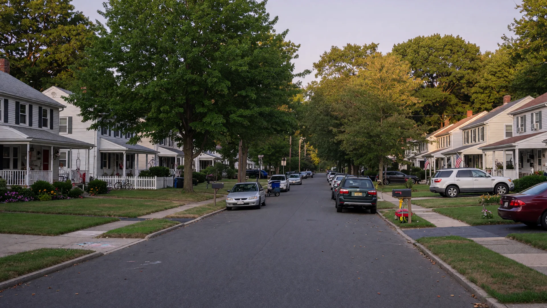 A quiet residential street in West Orange NJ with parked cars along the curb, mature trees, and single-family homes on a calm late-summer afternoon