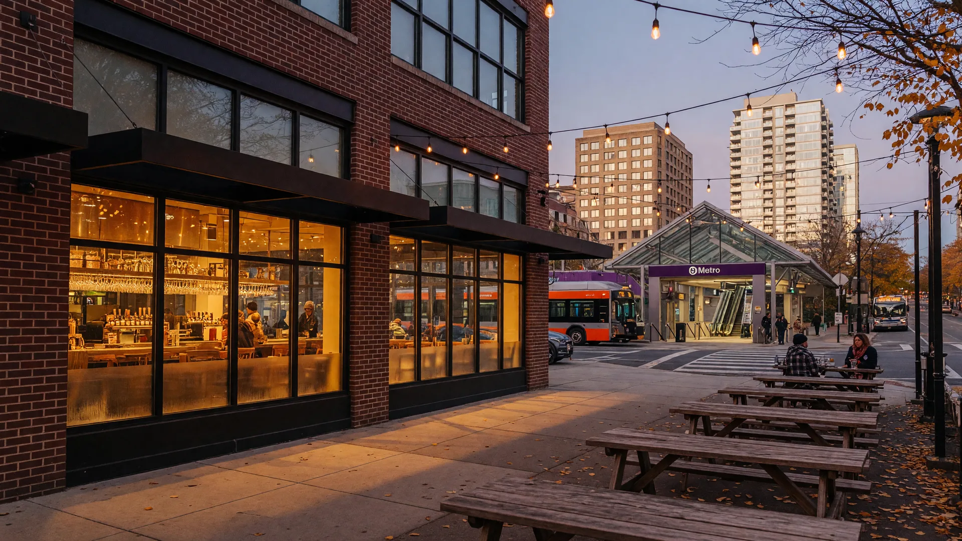 Modern brick-and-glass storefront of Silver Branch Brewing Company's biergarten in Downtown Silver Spring, Maryland at golden hour, with wooden tables and string lights on the sidewalk and the Silver Spring Metro station and downtown towers in the background