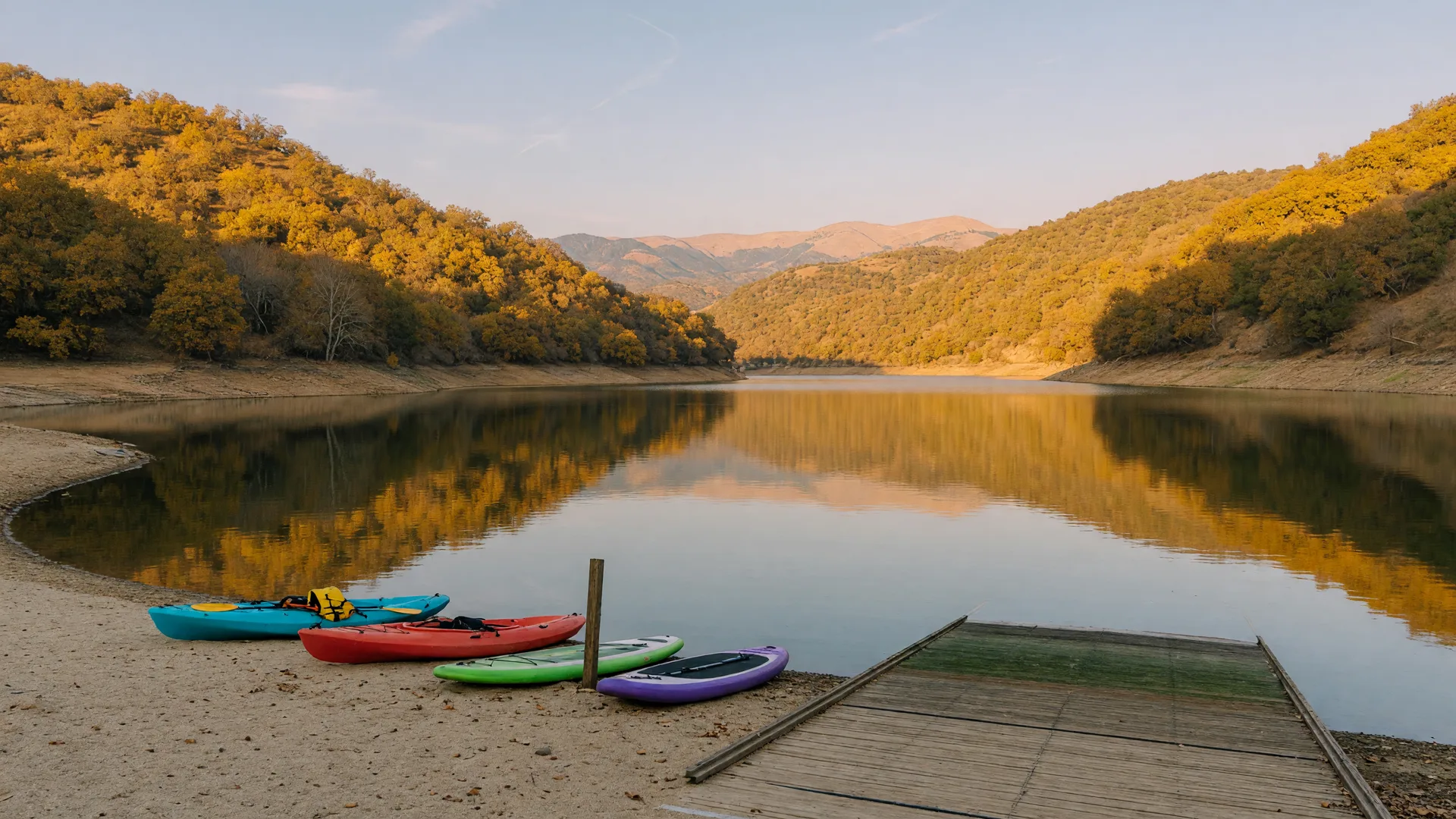 A calm Sierra-foothill reservoir in the Central Valley on a clear late-October afternoon, golden oak-covered hillsides, a sandy swim beach with colorful kayaks and paddleboards pulled up on the sand, glassy water reflecting the amber hills, and a wooden boat launch ramp