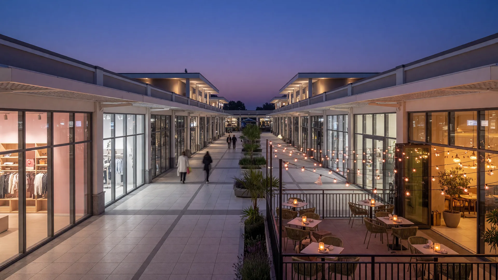 Bright modern shopping-center concourse at dusk with glass storefronts glowing warmly and a restaurant patio with string lights