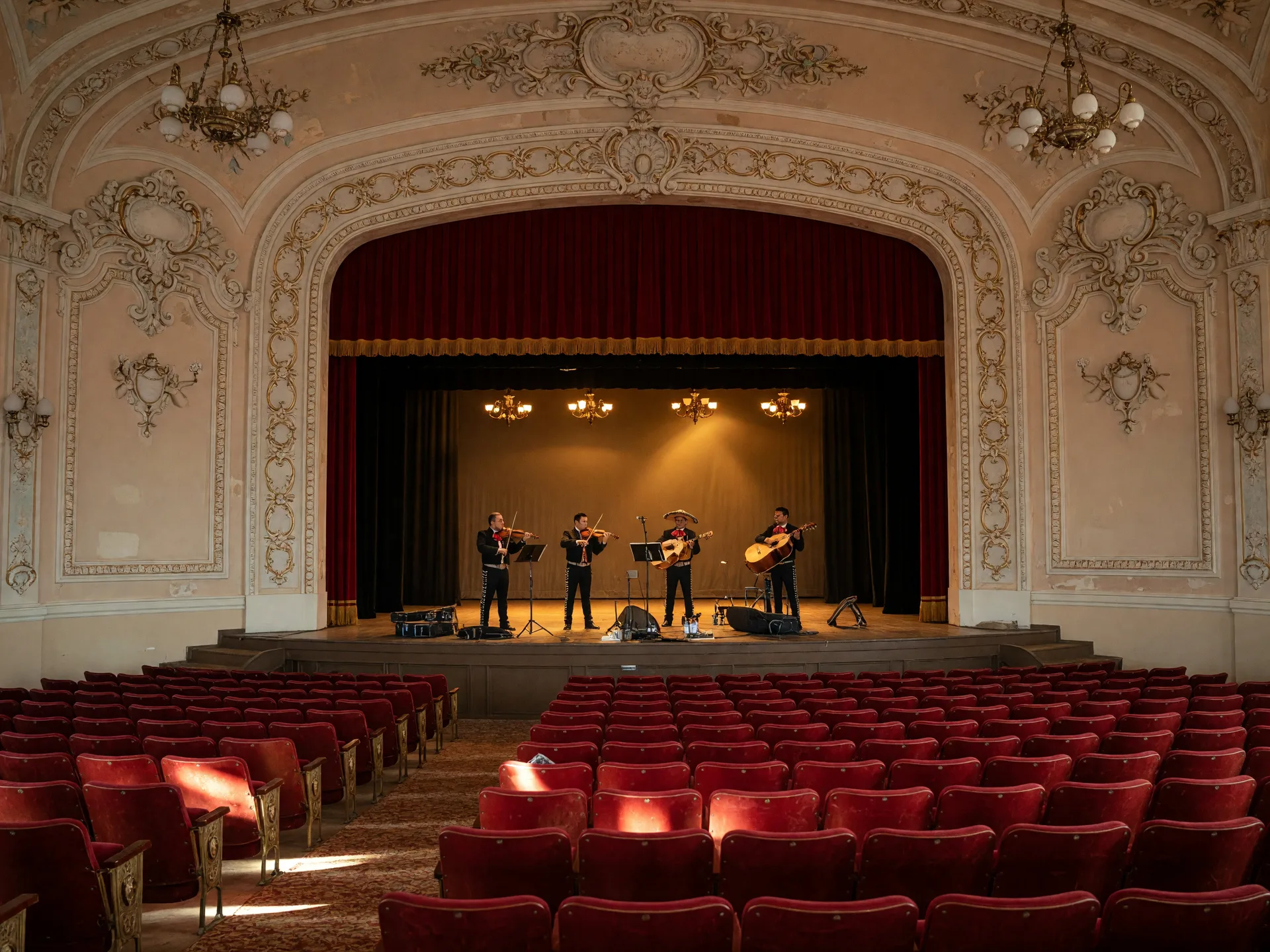 The restored interior of a historic Central Valley theater on a Friday evening, ornate plaster details and a deep red stage curtain, a mariachi band with violins and a vihuela setting up on stage, and red velvet seats in the warm glow of the house lights