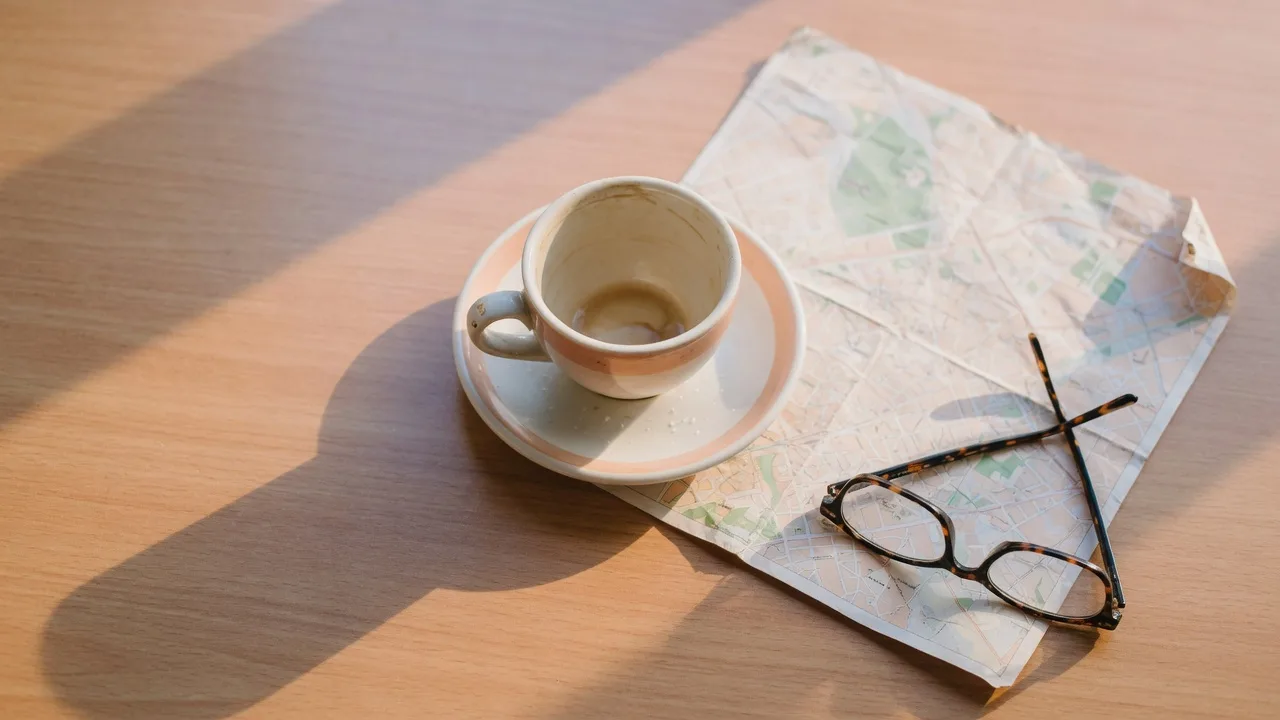 A ceramic coffee cup, a folded city map, and reading glasses on a warm wooden table