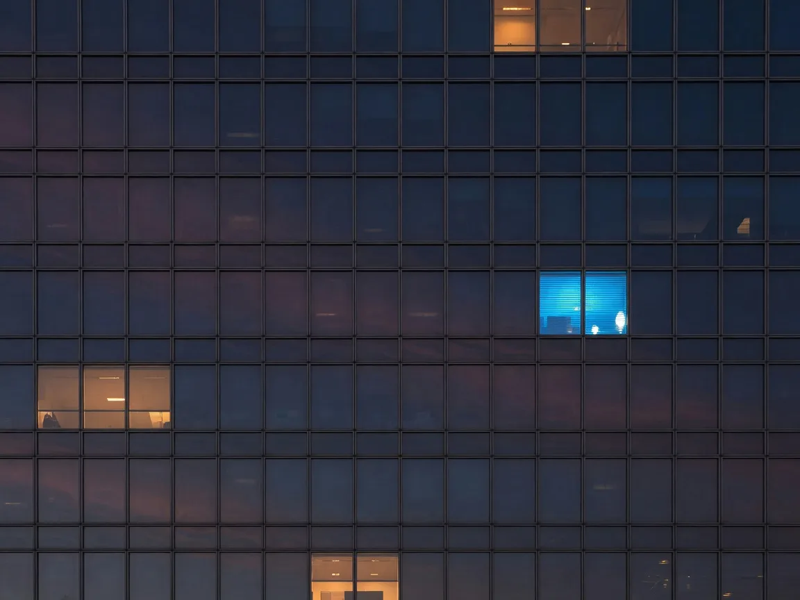 A glass facade at dusk with one electric-blue panel among warm-lit windows