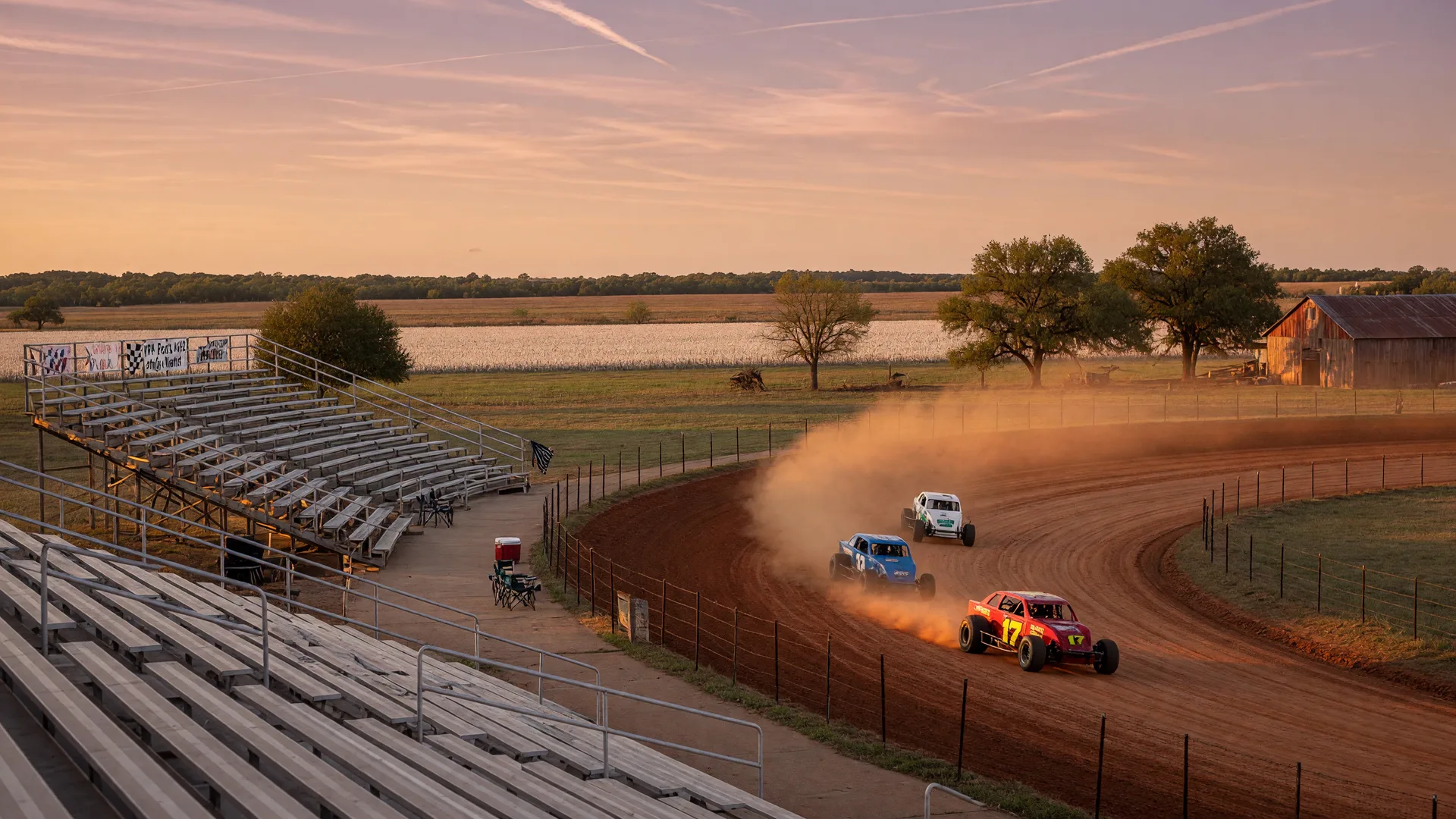 Dirt track race cars kicking up dust on a red clay oval in rural Northeast Texas at golden hour