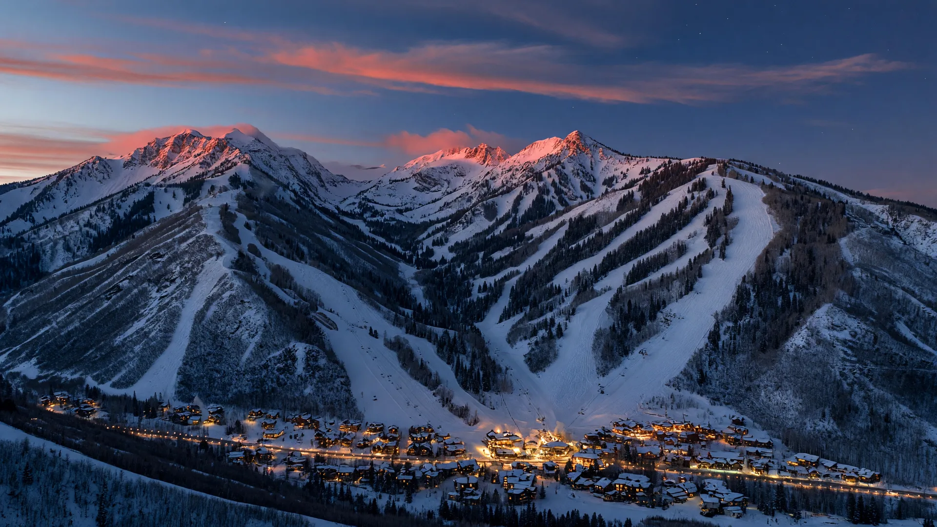The Wasatch Mountains at winter twilight, groomed ski runs dropping from a dark ridgeline toward a glowing resort village far below