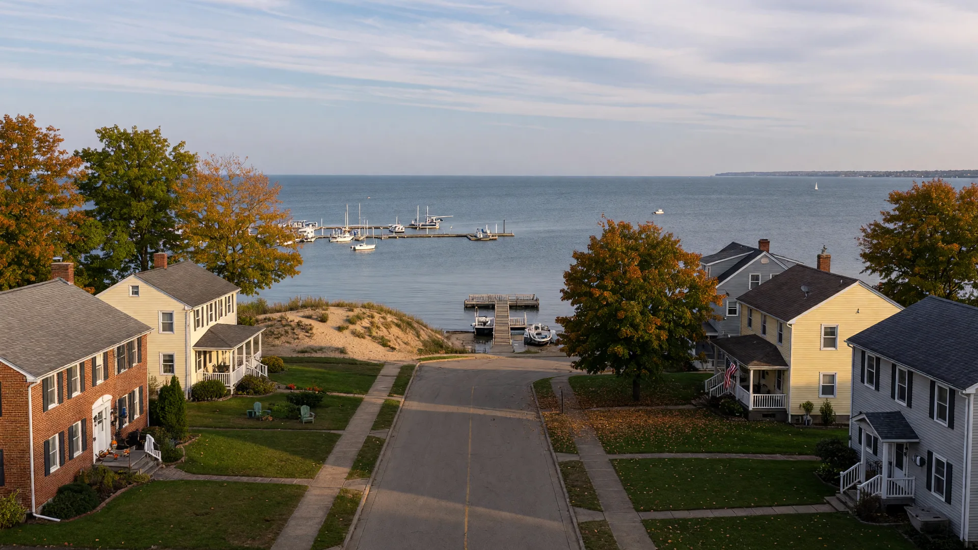 A Lake Erie shoreline street in Madison, Ohio in early October with brick and colonial homes under maples turning orange and gold, a sandy bluff and marina sailboats on the lake beyond